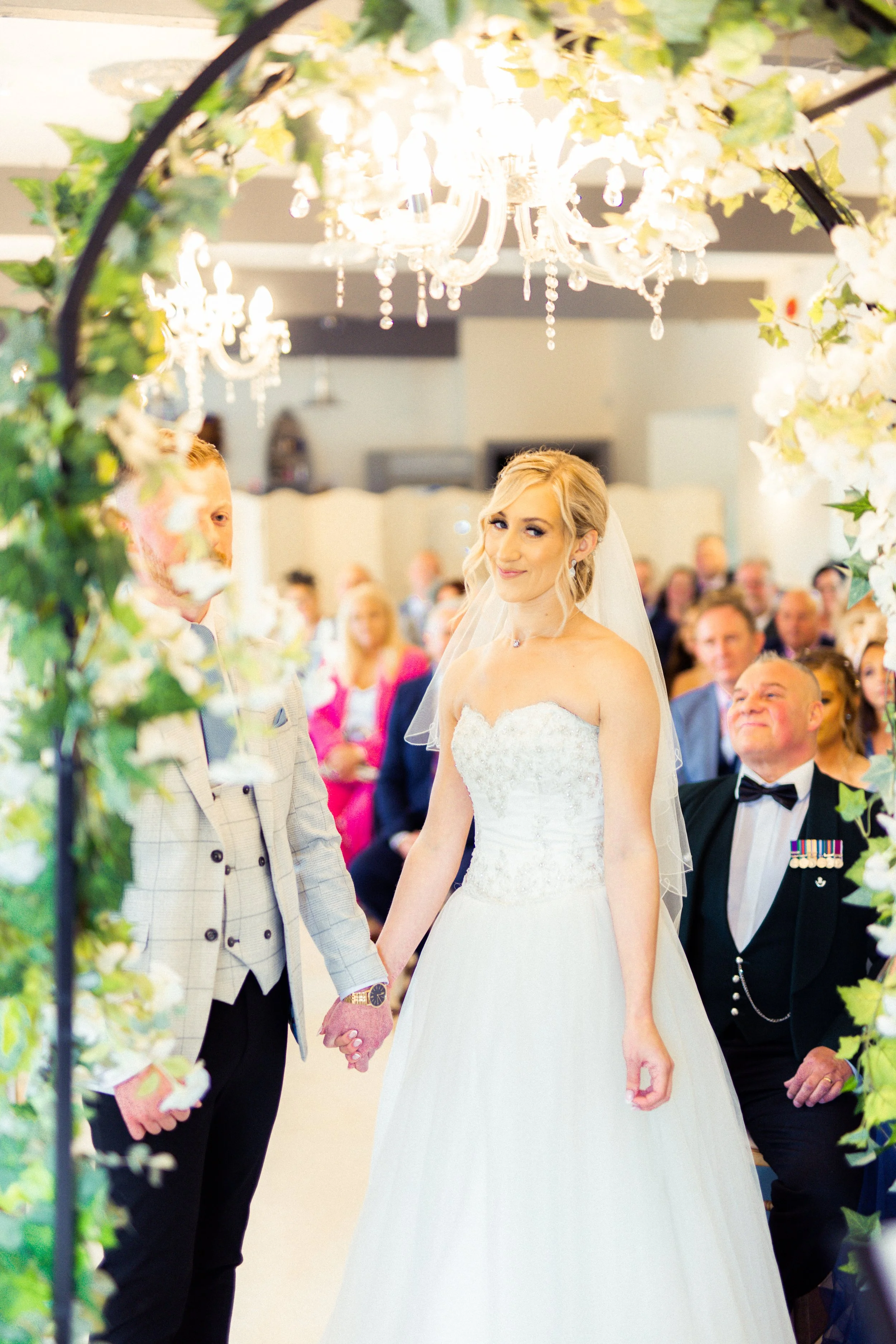 Bride and groom holding hands during their wedding ceremony, surrounded by friends and family, with floral decorations and chandelier lighting.