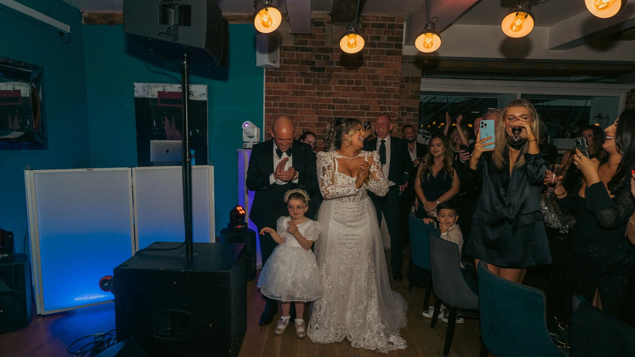 People celebrating at a wedding reception, with a bride in a lace wedding dress, a man in a black suit, a young girl in a white dress, and guests taking photos and videos in a decorated indoor venue with brick walls and warm lighting.