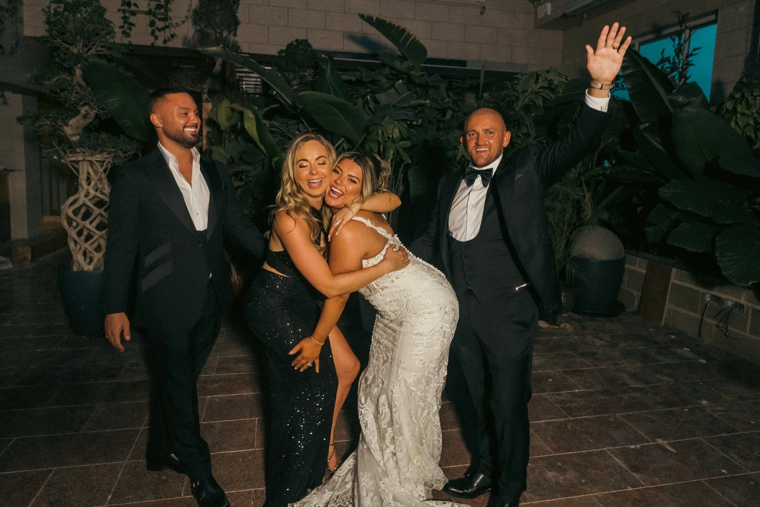 Group of five people celebrating at a wedding reception, with two women in the center, one in a wedding dress and the other in a black gown, hugging and smiling, surrounded by men in tuxedos, all appearing joyful.
