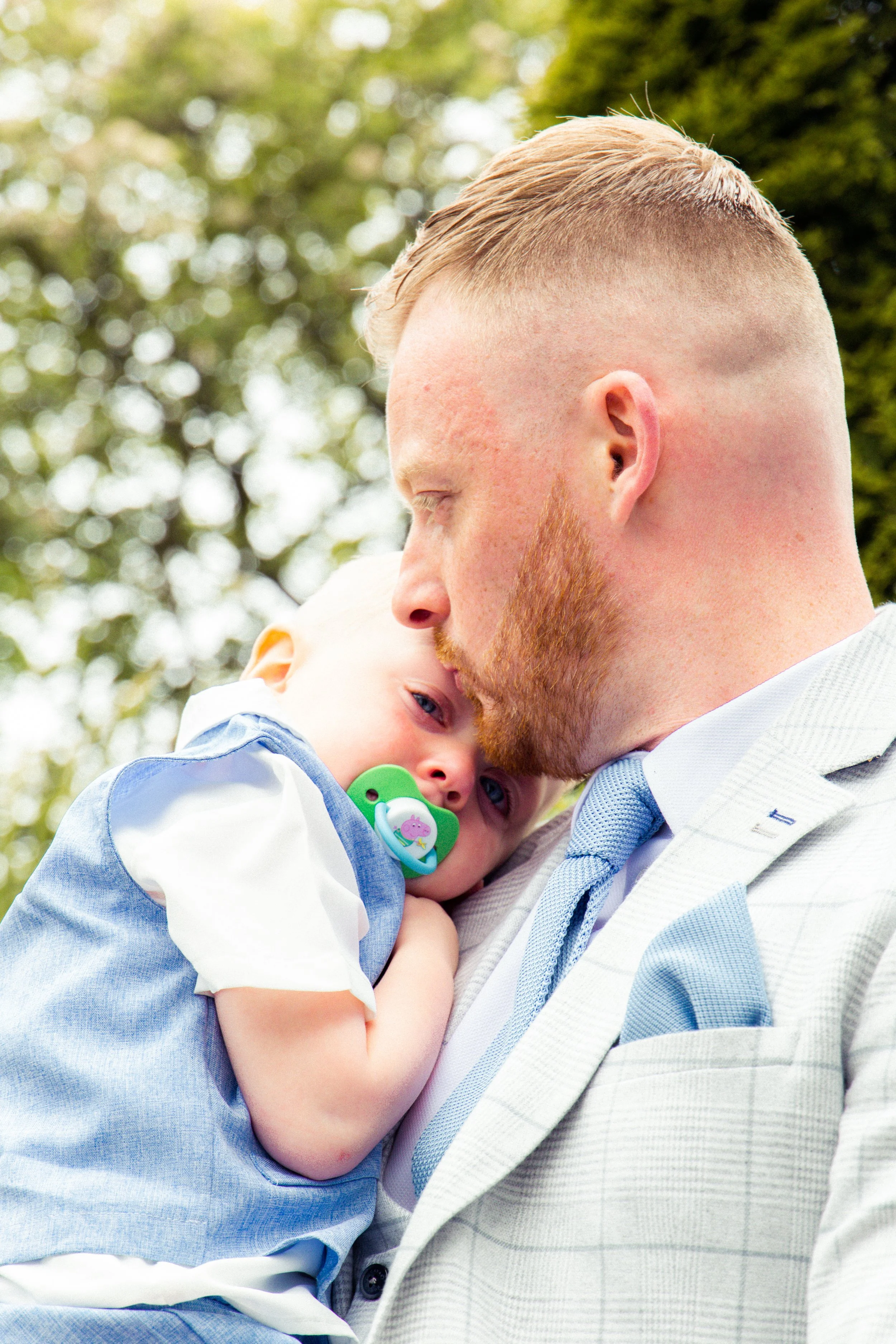 A man dressed in a light suit holding a young child with a pacifier, outdoors with green trees in the background.