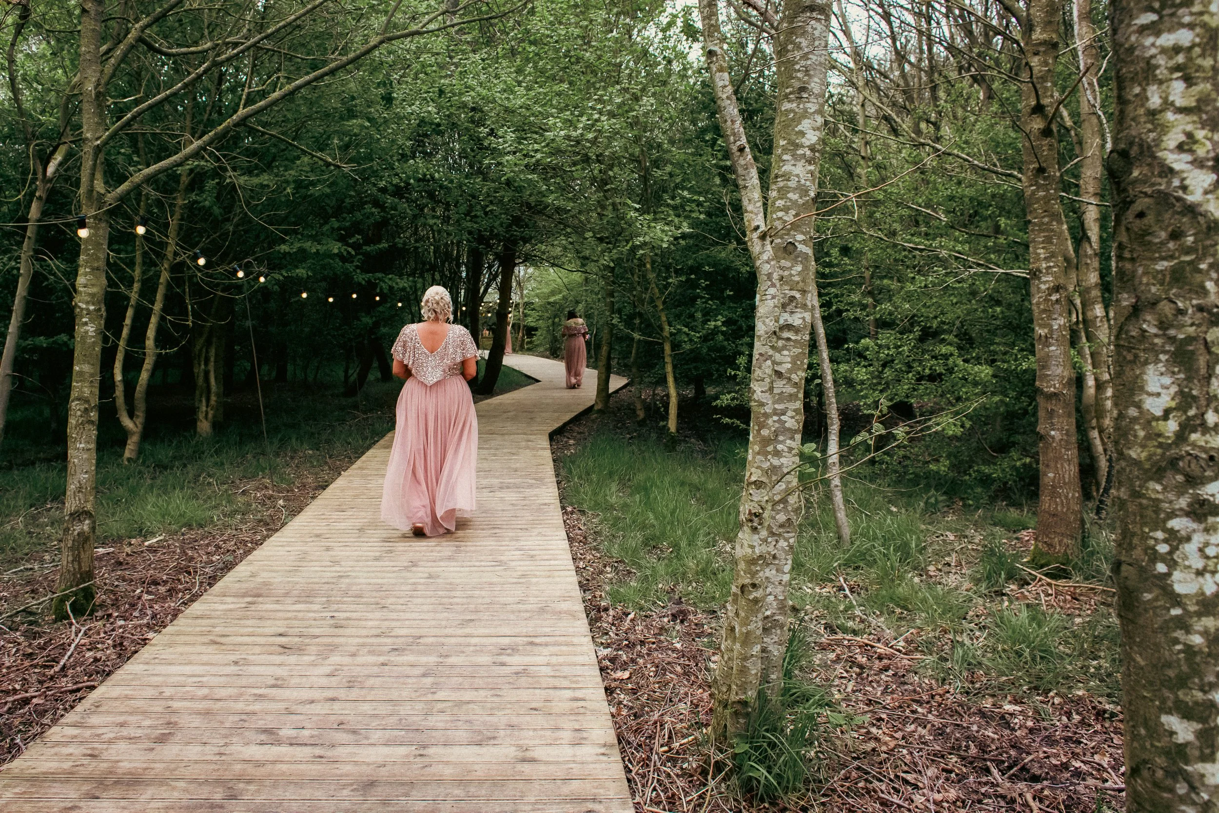 A woman in a long pink dress walking on a wooden pathway through a green forest with trees on either side.