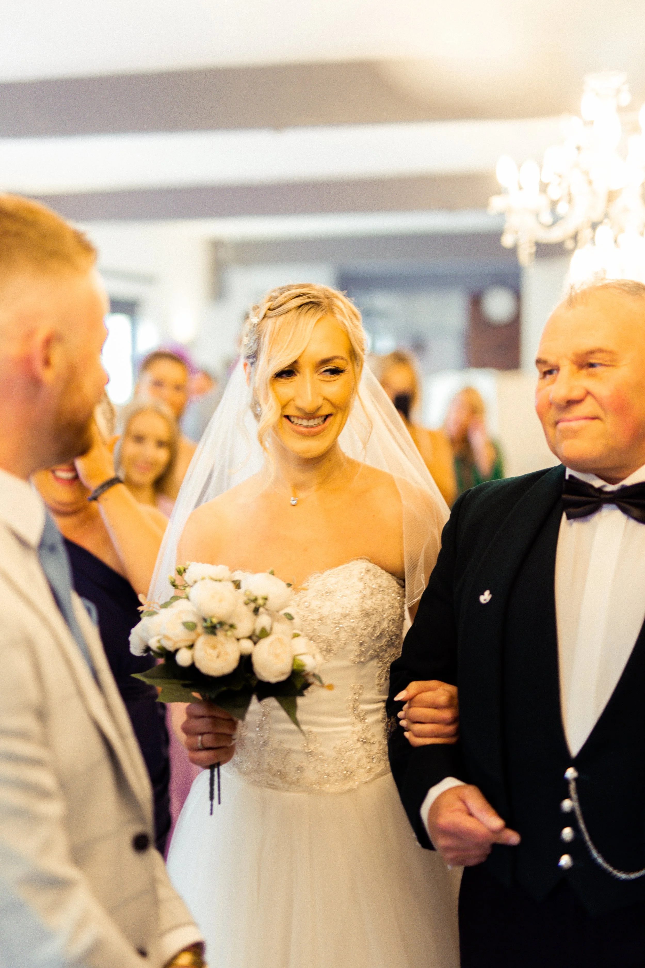 Bride smiling, holding a bouquet of white flowers, standing between two men in formal attire during a wedding ceremony.