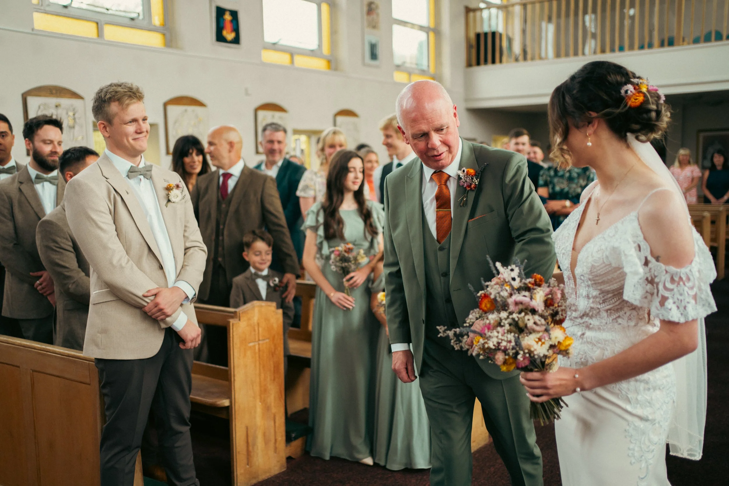 A bride in a white lace wedding dress holding a bouquet of dried flowers, talking with an older man in a green suit, inside a church during a wedding ceremony. Guests are standing and smiling in the background.