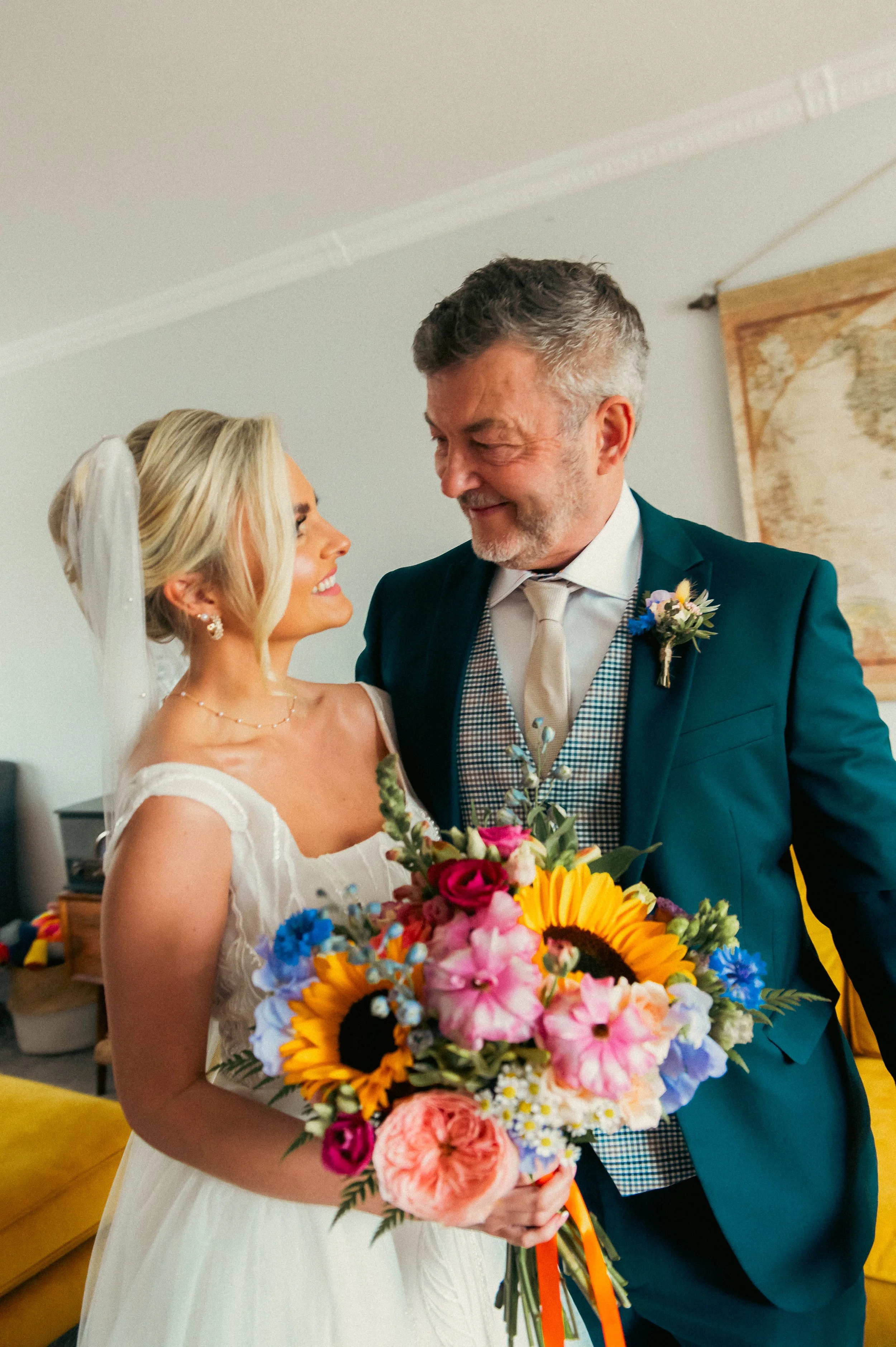A bride and an older man smiling at each other indoors. The bride is holding a large bouquet of colorful flowers, and is wearing a wedding dress with a veil. The older man is wearing a teal suit jacket, a white shirt, and a tie, with a small floral b