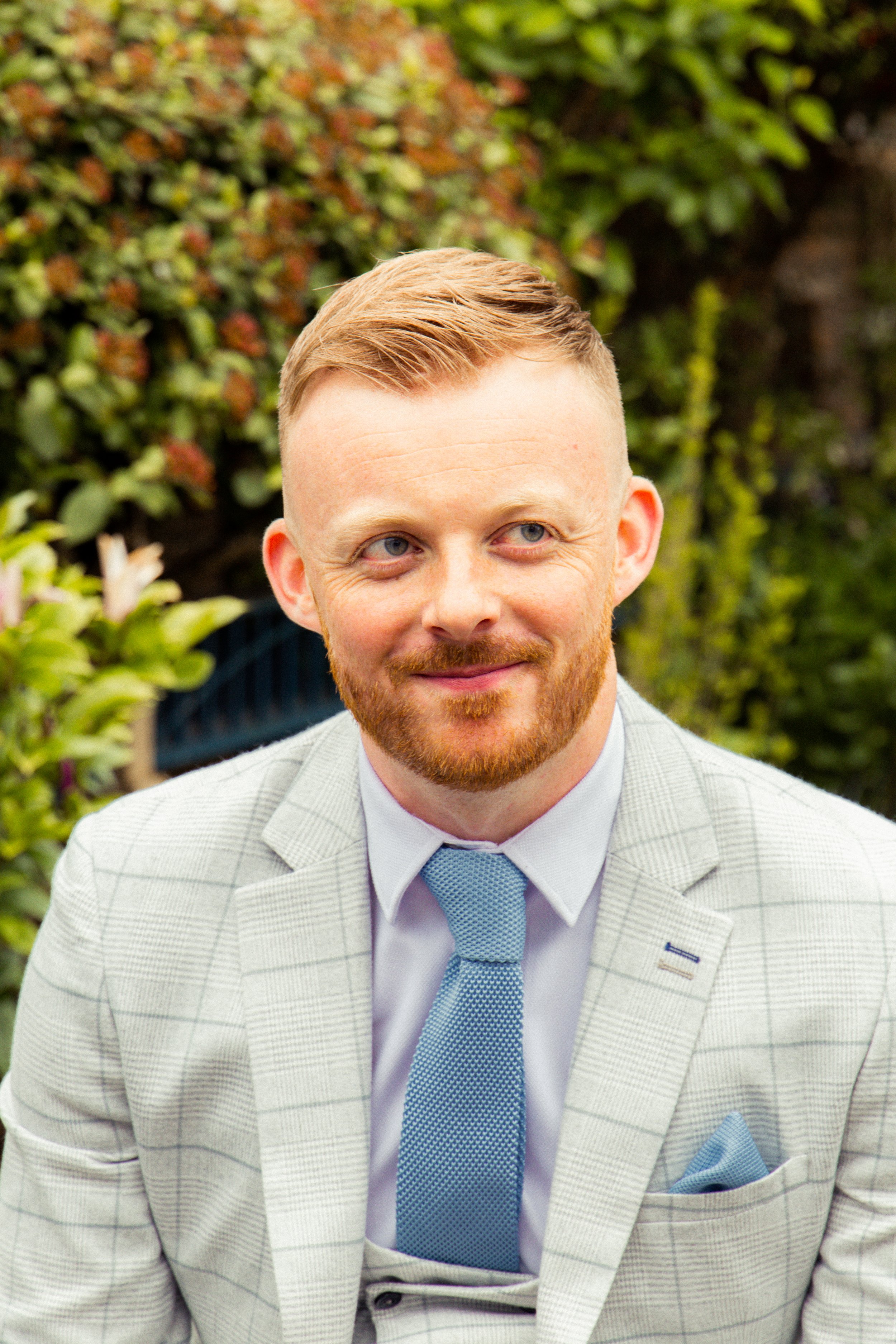 A man with ginger hair, a beard, and blue eyes wearing a light gray checkered suit, light blue shirt, and matching blue tie, standing outdoors with greenery in the background.