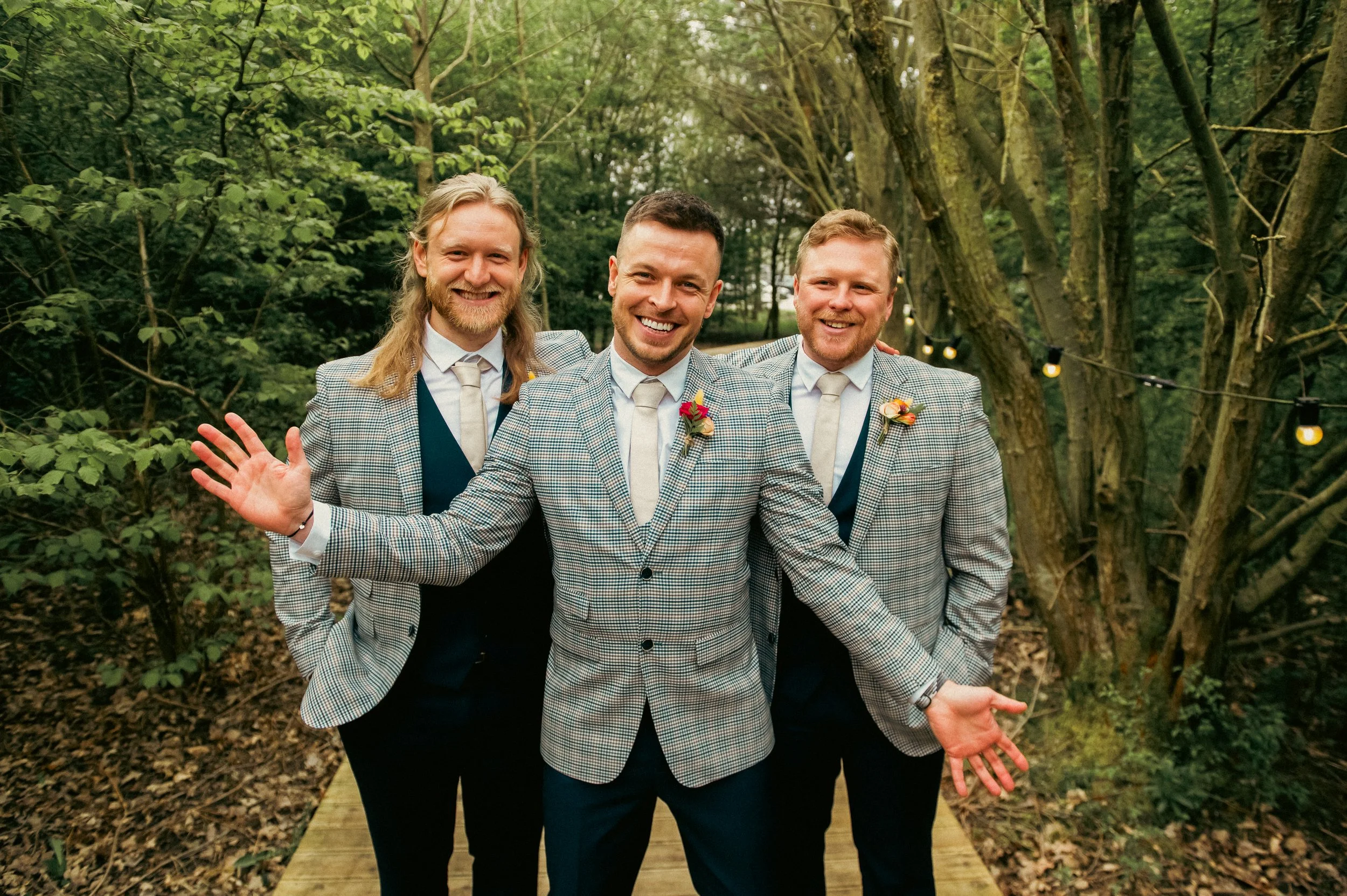 Four men dressed in suits and ties, standing outdoors on a wooden path with trees behind them, smiling and posing for the camera.