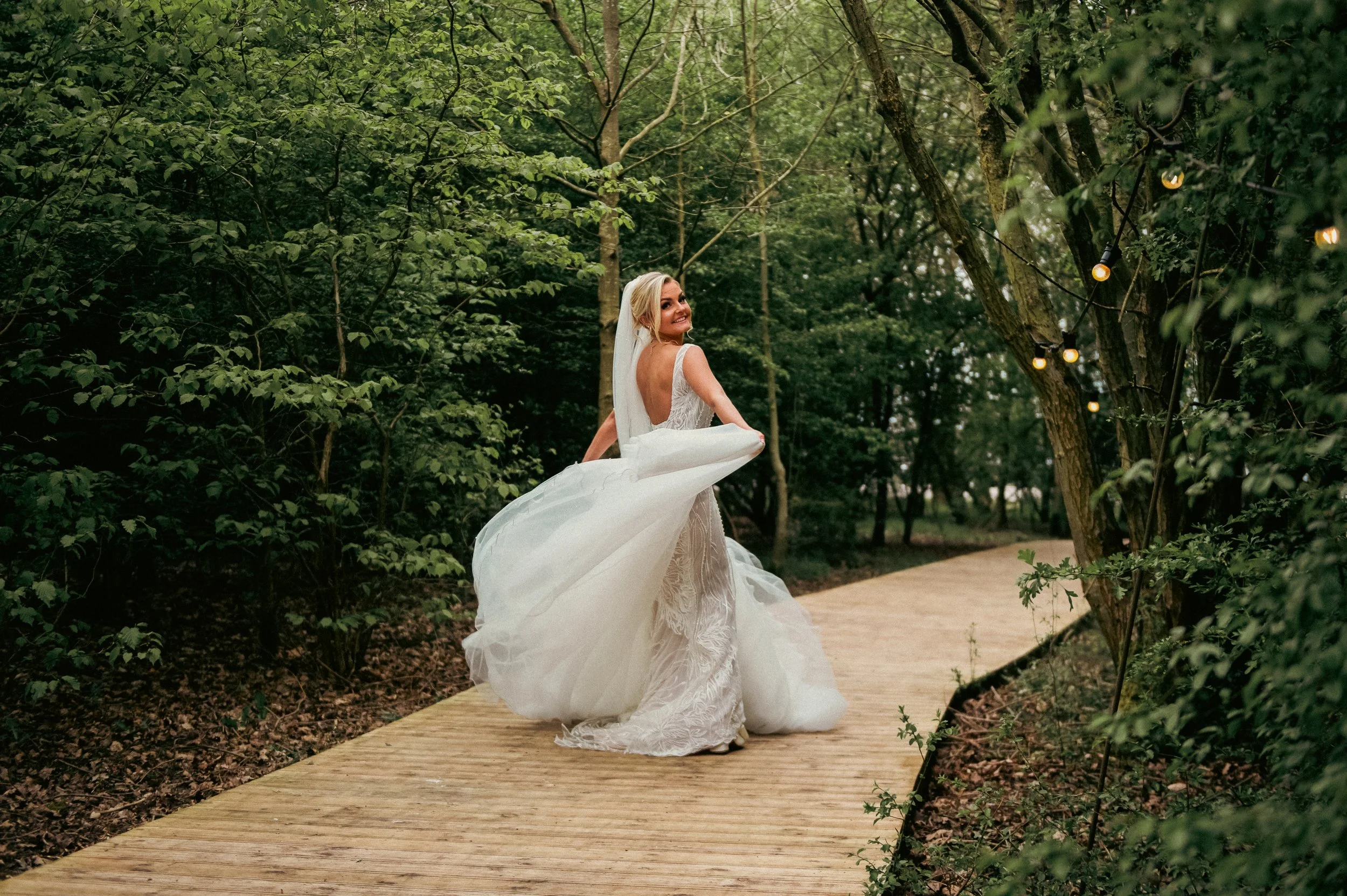 A bride in a wedding dress standing on a wooden pathway in a lush green forest, smiling and holding her dress