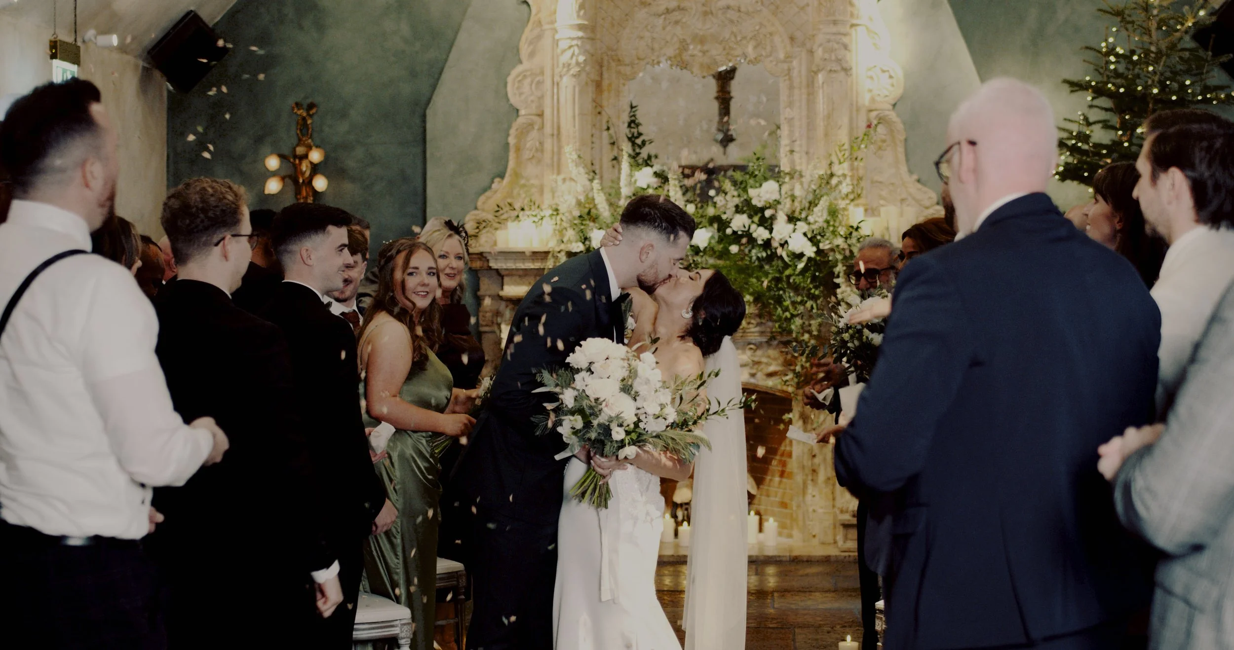 A wedding ceremony with a bride and groom sharing a kiss, surrounded by guests in an elegant indoor setting with floral arrangements and candles.