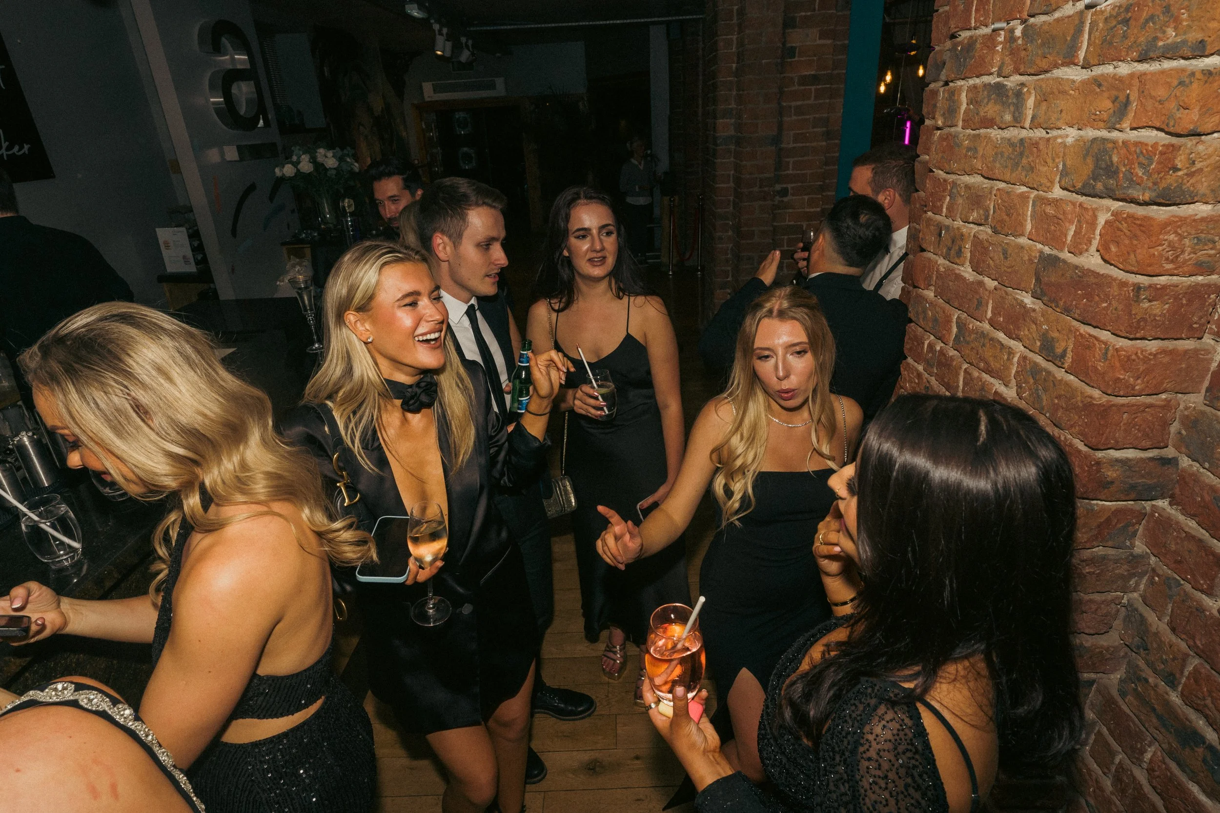 Group of people at a party in a brick-walled venue, holding drinks and engaging in conversation.