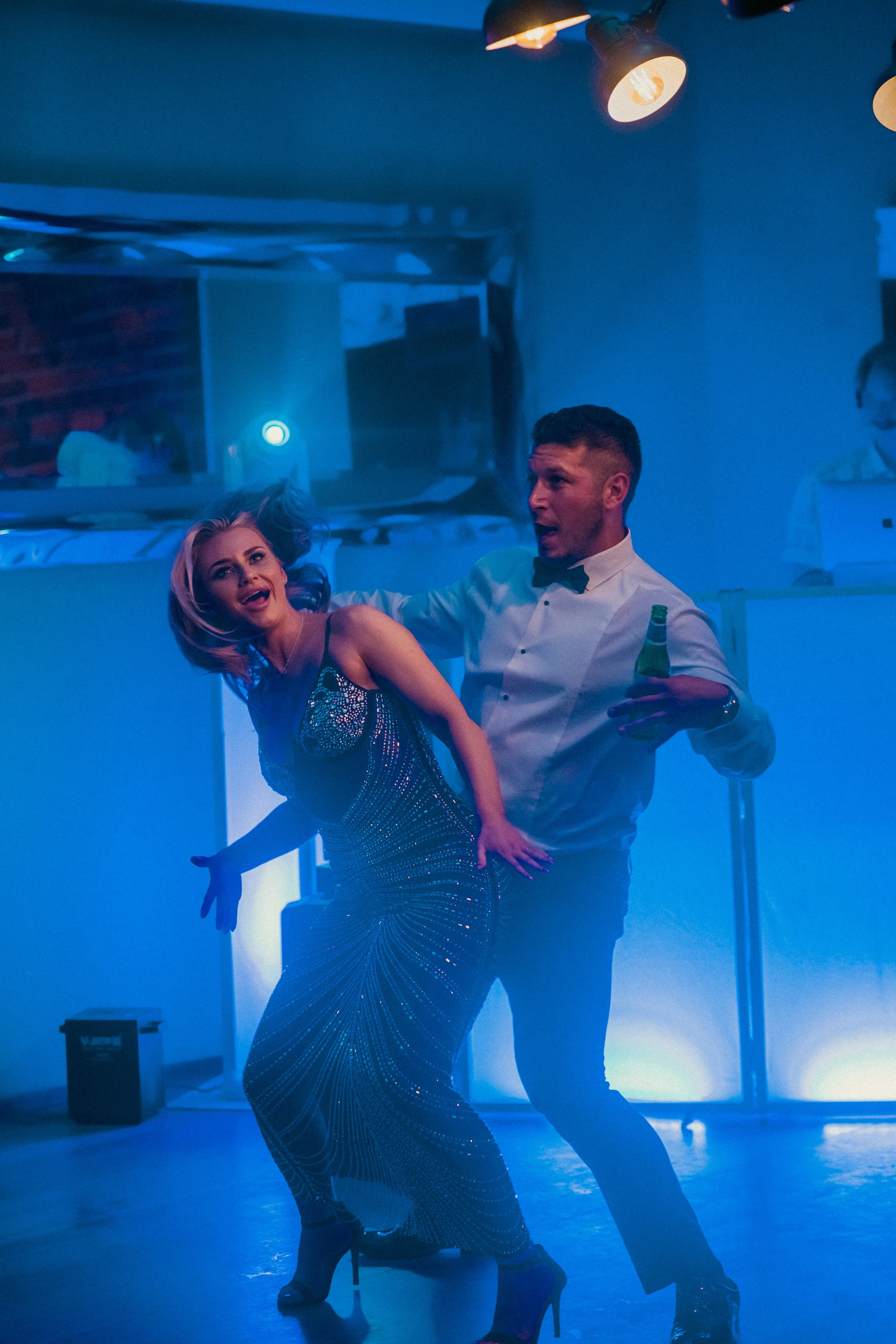 A woman in a sparkly dress and high heels dancing with a man in a white shirt and bow tie at a party or nightclub. The man is holding a green bottle, and there are DJ or lighting equipment and colorful stage lights in the background.
