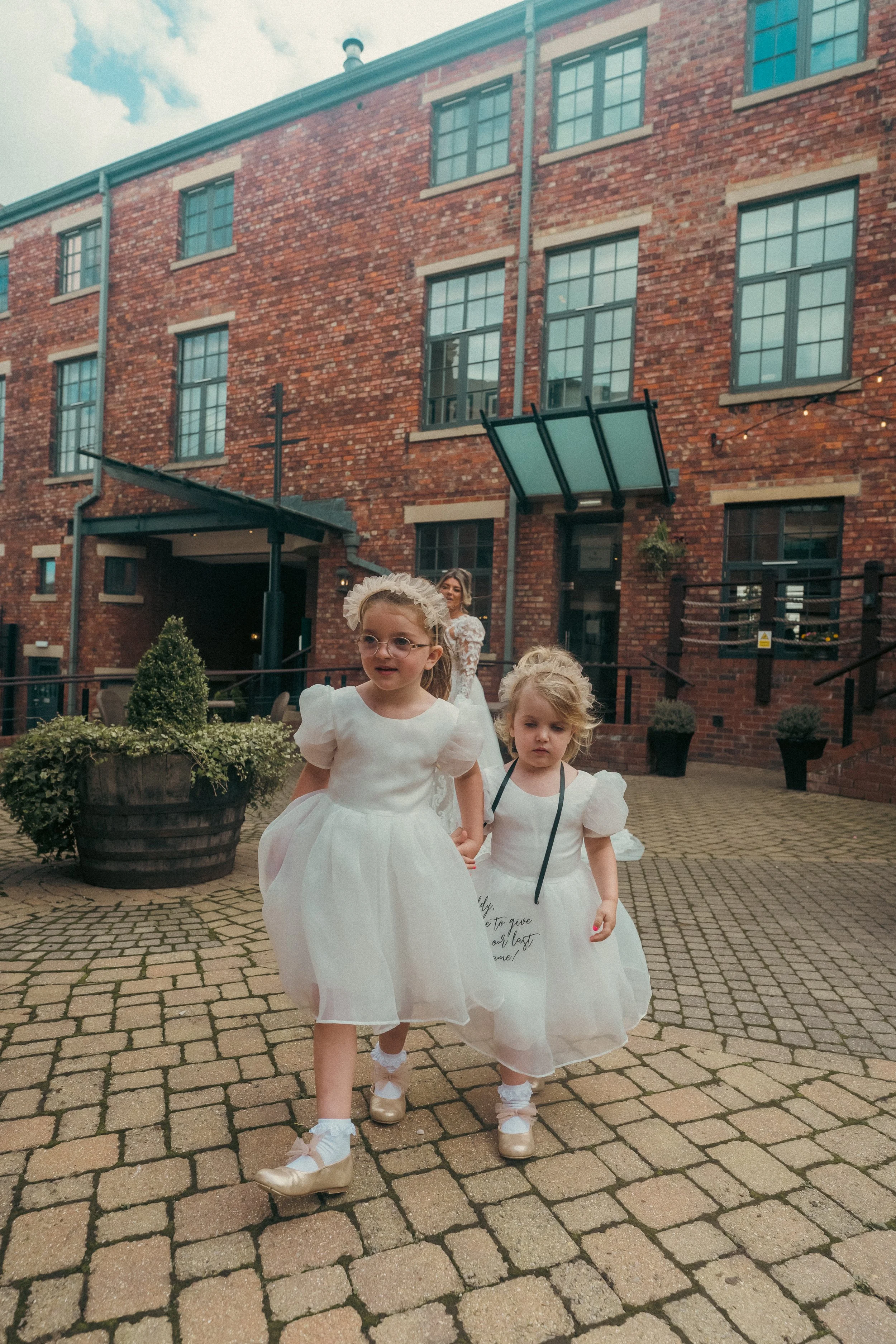 Two young girls in white dresses walking outside on a brick patio, with a woman in a white dress behind them, near a brick building with large windows and potted plants.
