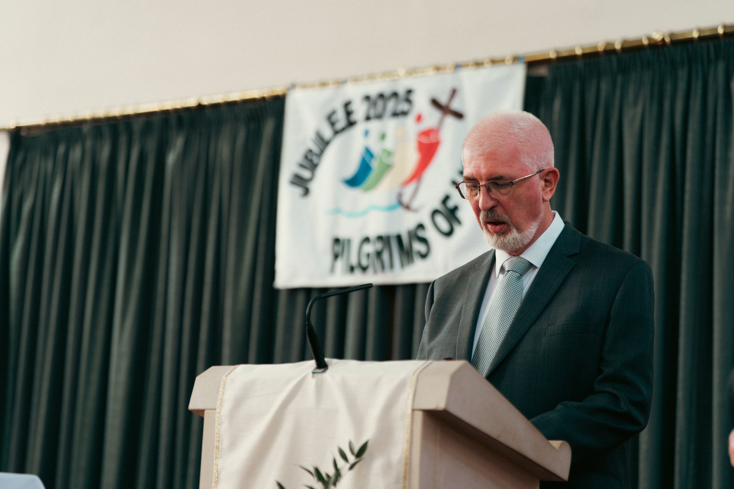A man with glasses and a white beard dressed in a suit, standing at a podium with a white cloth, speaking into a microphone at an event with a green curtain backdrop and a banner that reads 'Journey 2025 Pilgrims of' with a design of three stylized f