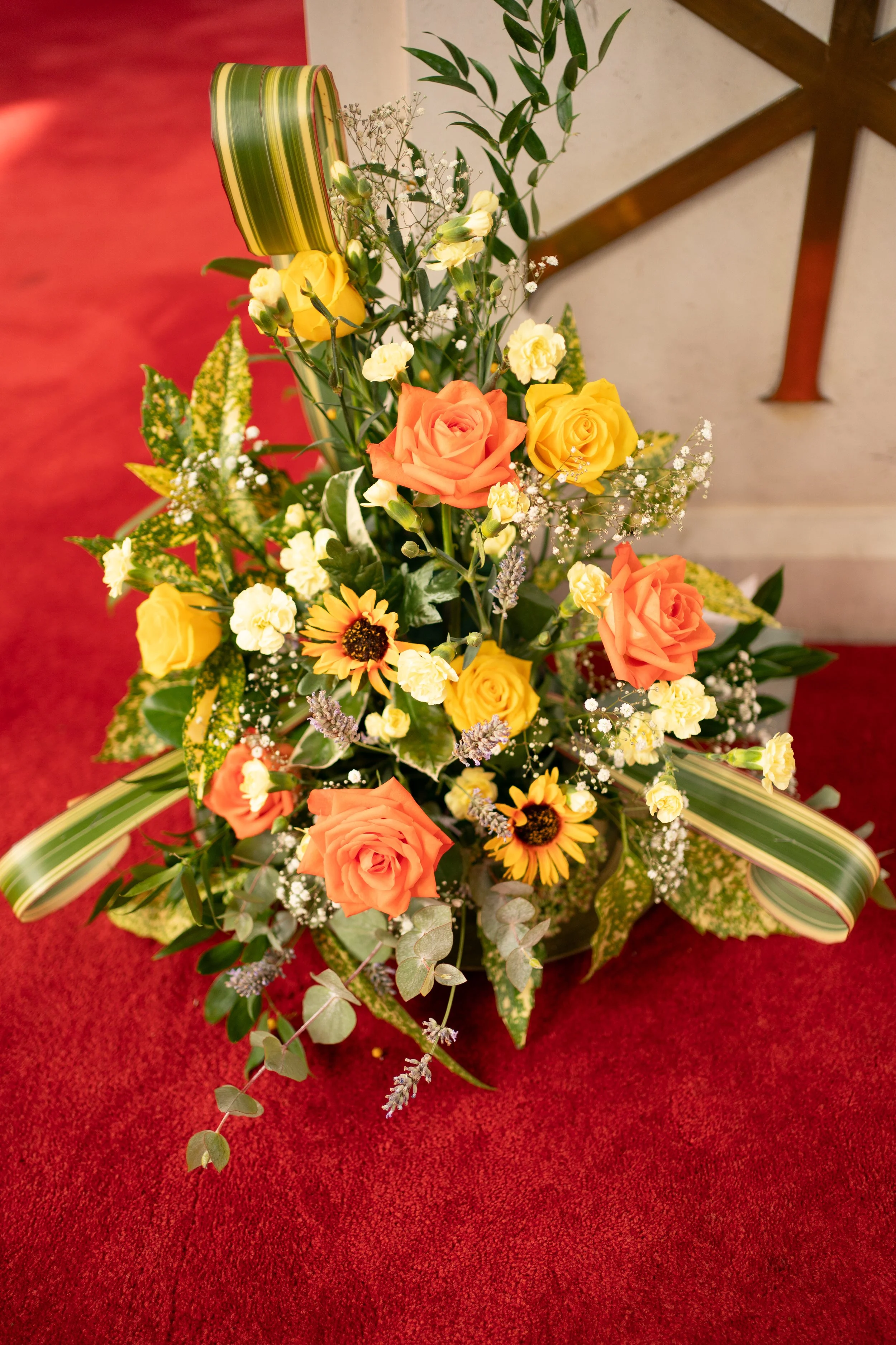 A colorful flower arrangement with roses, sunflowers, and greenery, placed on a red carpeted surface near a wall and a wooden clock.
