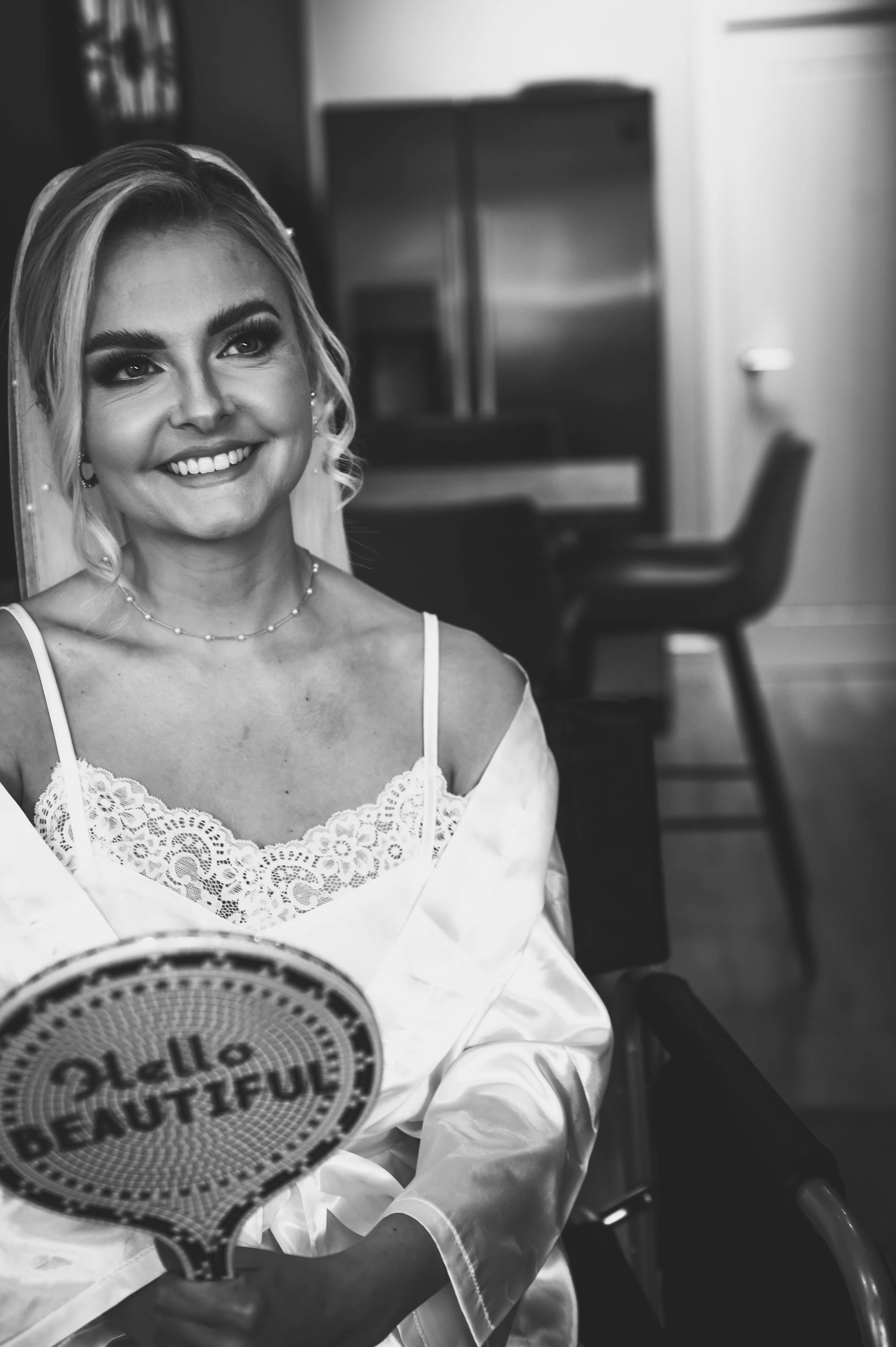 A woman with blonde hair and makeup, dressed in a lacy camisole and satin robe, smiling and sitting in a wheelchair, holding a sign that says 'Hello Beautiful' in a room with chairs and a cabinet in the background.
