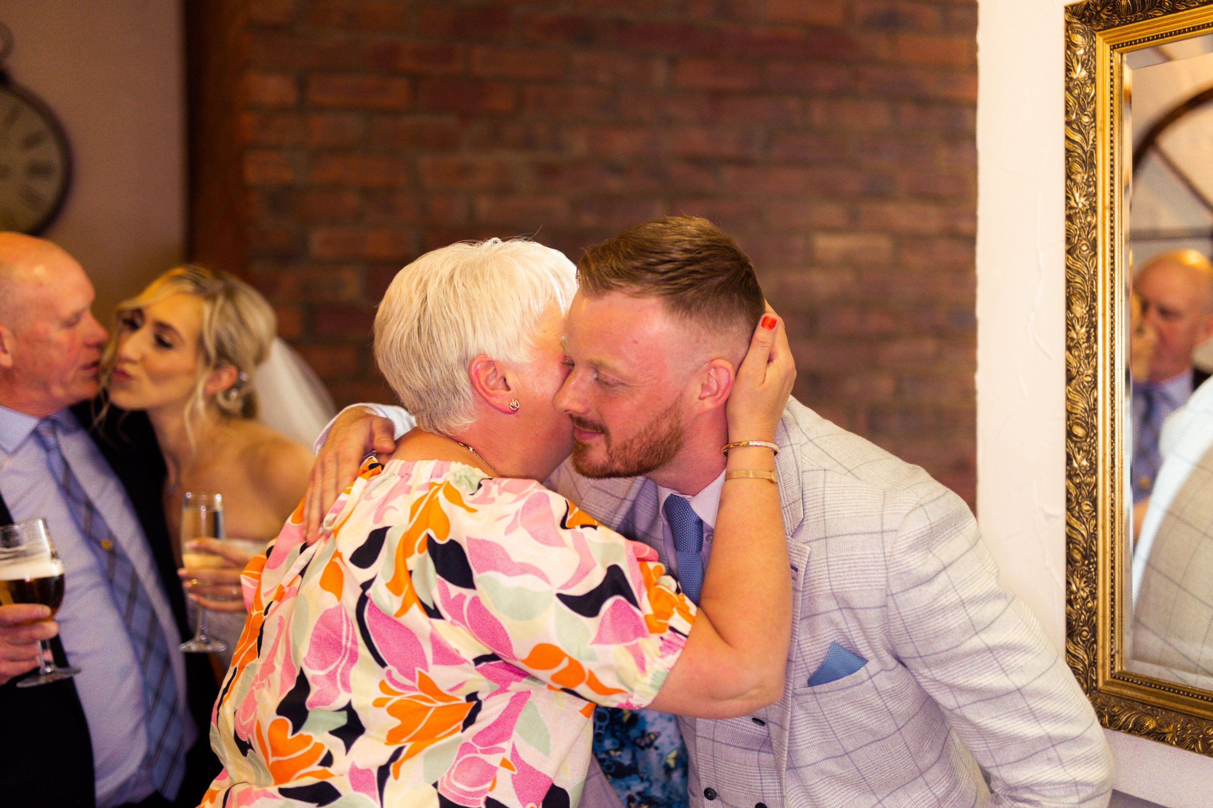 A woman with short blonde hair wearing a colorful floral dress is giving a kiss on the cheek to a young man with short brown hair and a beard, dressed in a light-colored checkered suit. They are indoors at a celebration with other people in the backg
