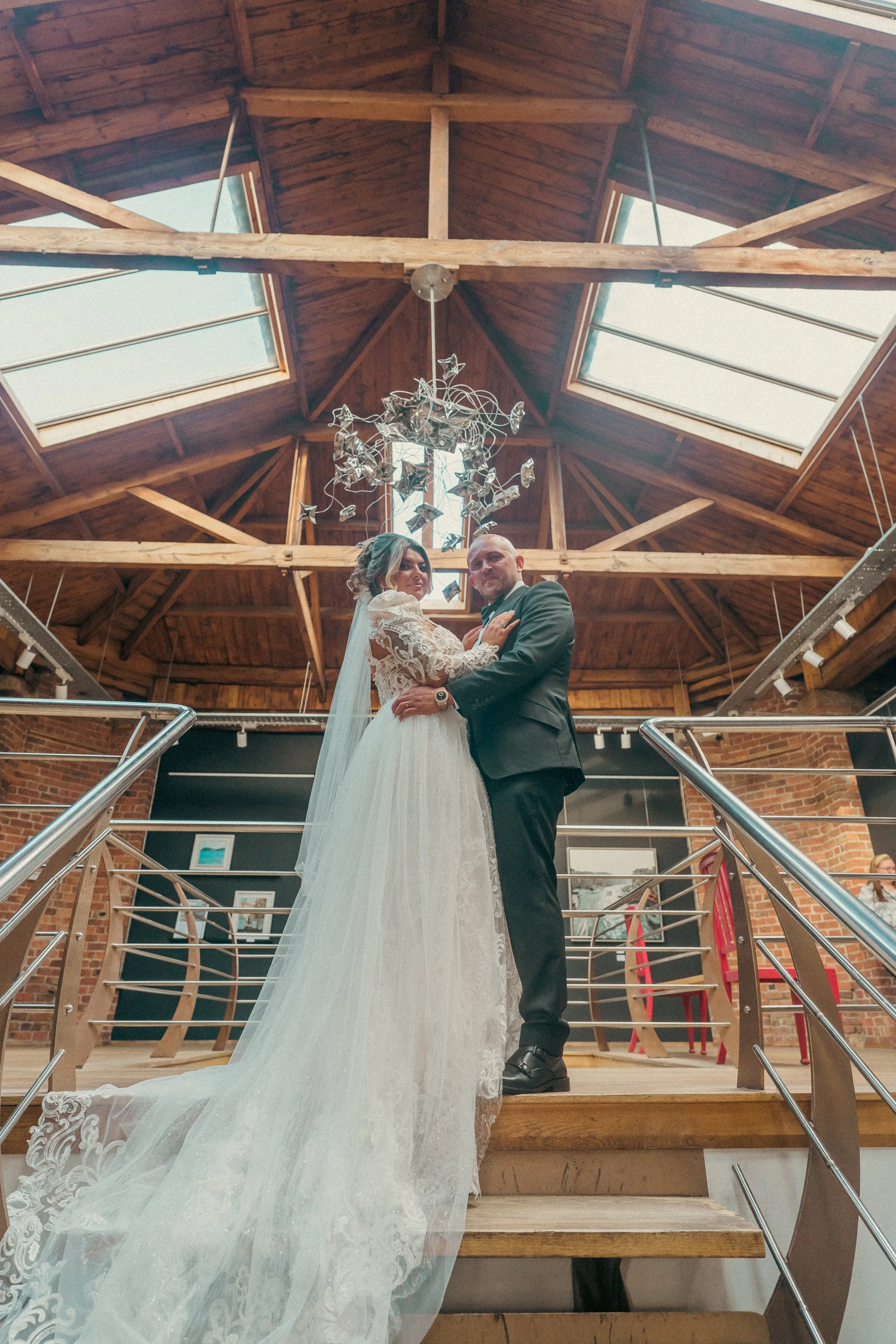 A bride and groom standing closely together on a wooden staircase, indoors, with a wooden ceiling and skylights above. The bride is in a lace wedding gown with a long train, and the groom is in a dark suit. They are looking at the camera.