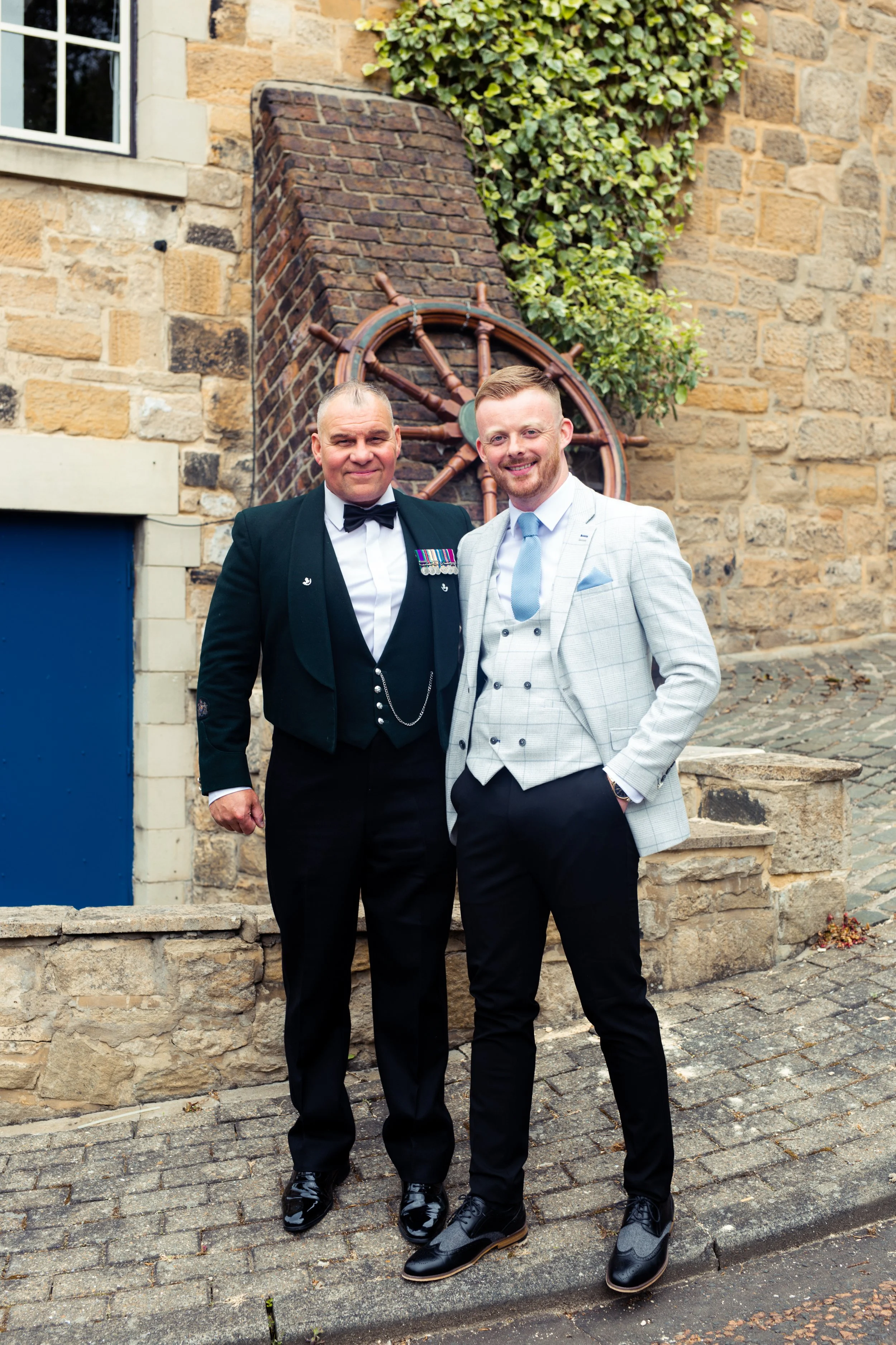 Two men dressed in tuxedos standing outdoors in front of a brick wall with a ship wheel decoration and greenery.