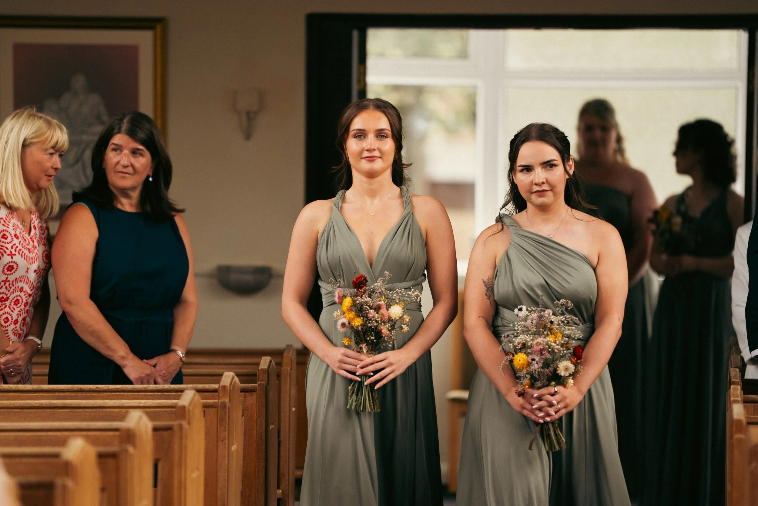 Two bridesmaids in green dresses holding bouquets standing in a church, with guests seated behind and to the left.