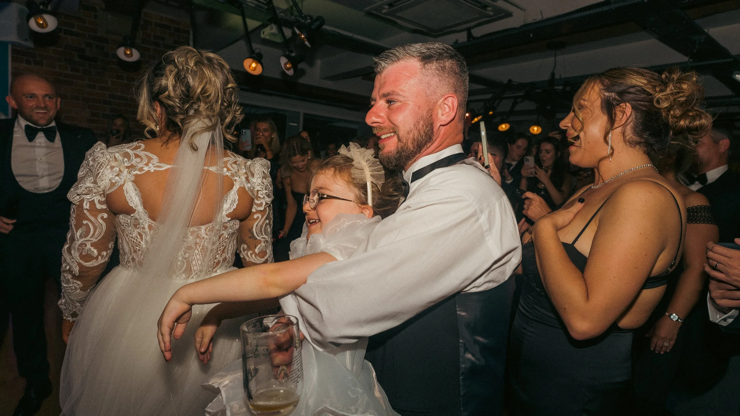 A group of people dancing and celebrating at a wedding reception, with a bride in a lace wedding dress, a man in a tuxedo, and a young girl in a white dress with glasses, smiling and enjoying the moment.