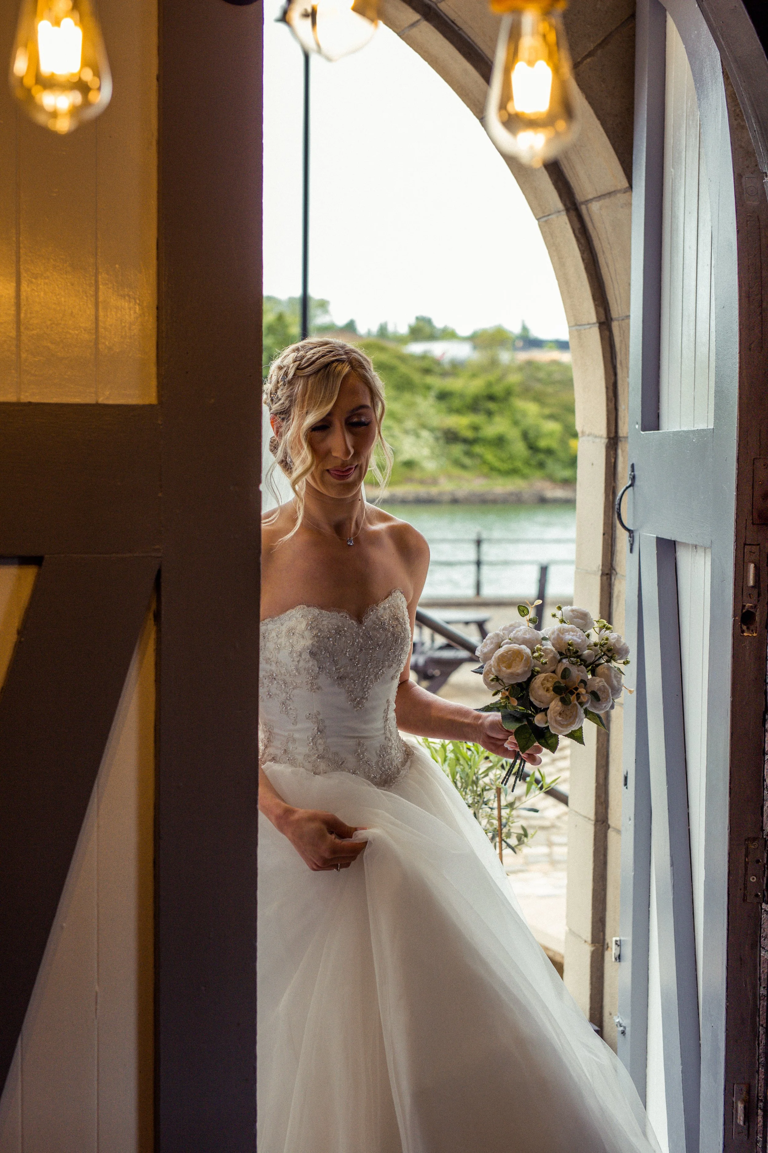 Bride in a strapless wedding dress holding a bouquet of white flowers, standing in the doorway of a building with a riverside view in the background.