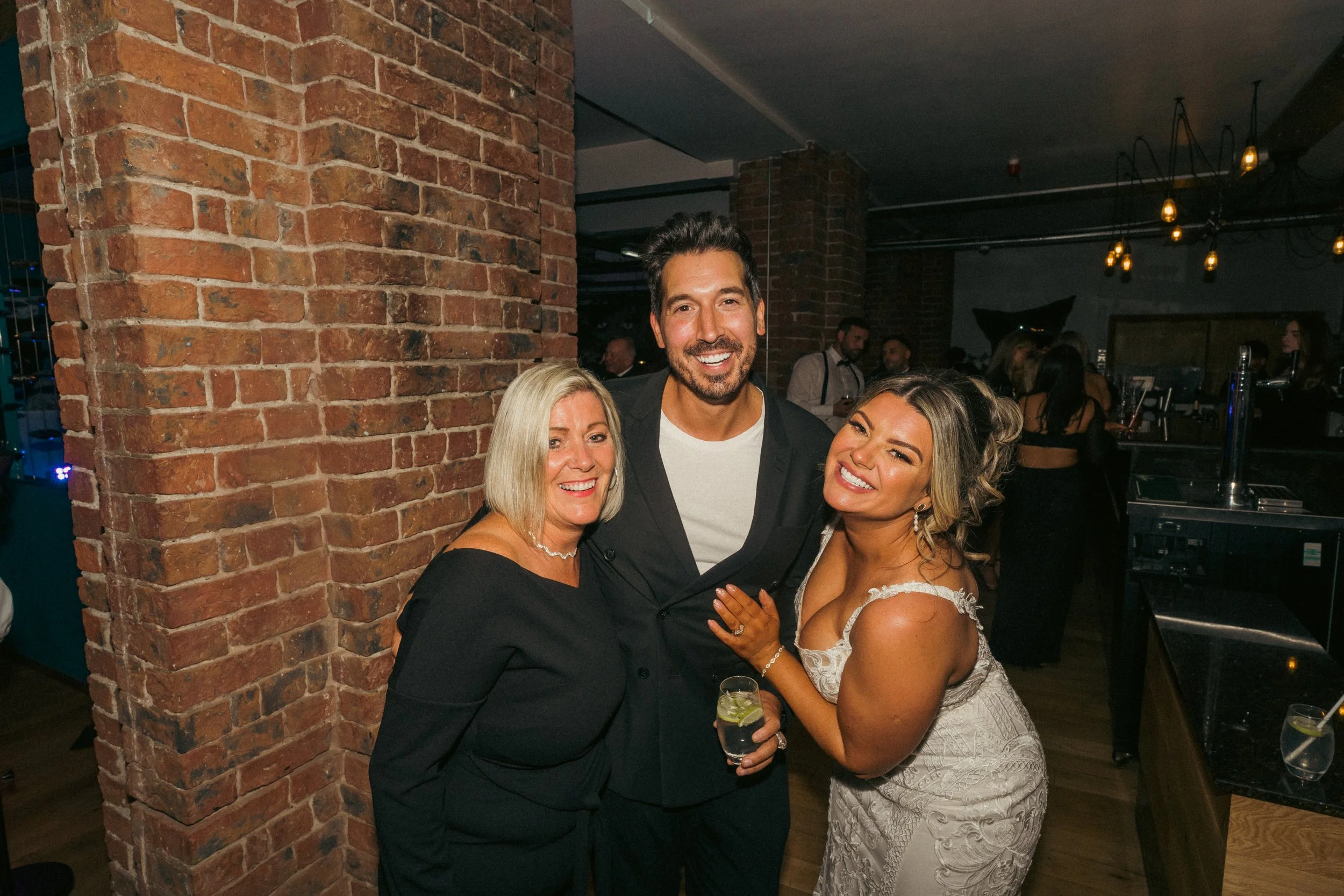 Three people smiling and posing for a photo at a social event, standing close together near a brick wall in a dimly lit venue, with other guests in the background.