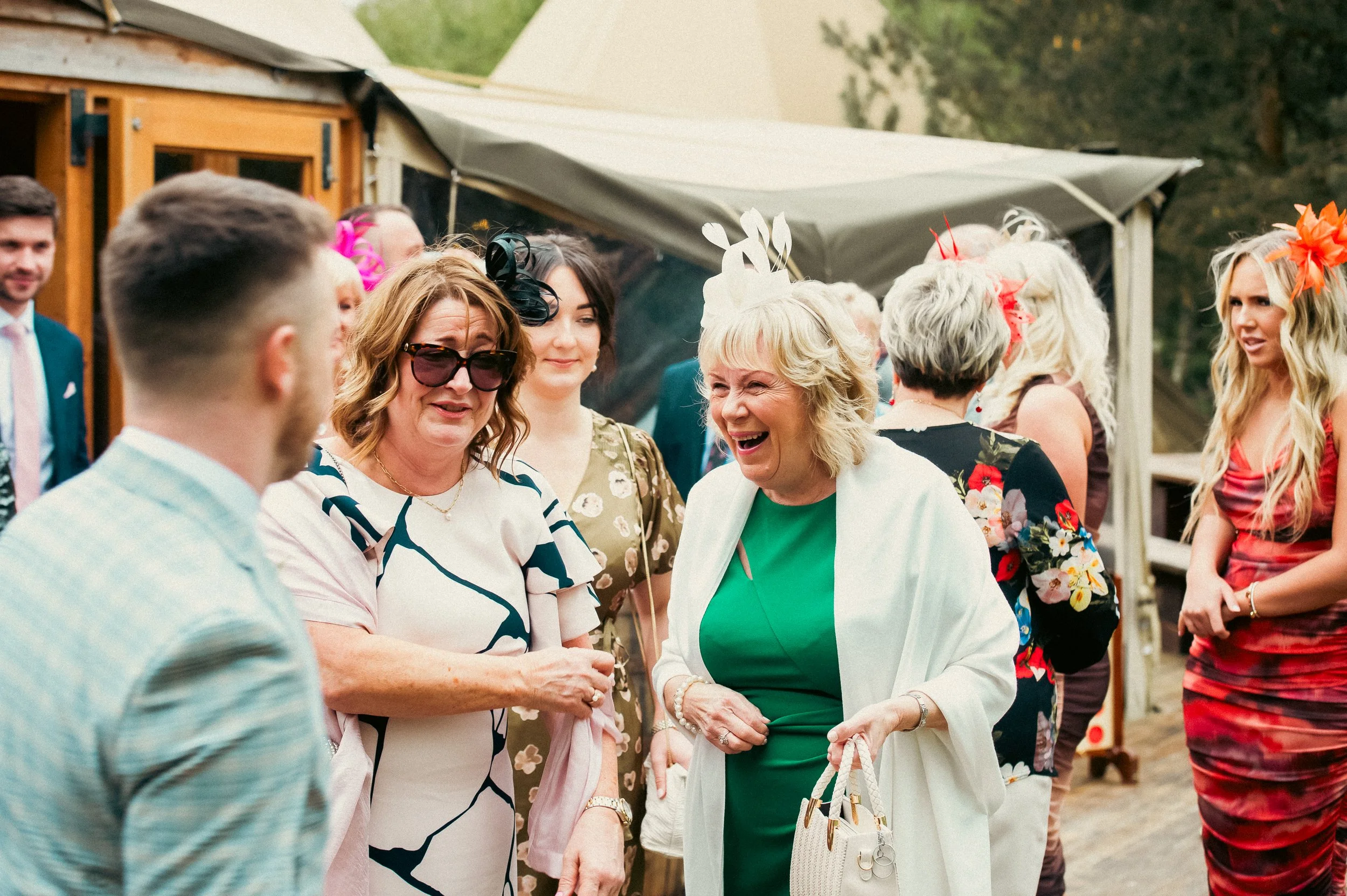A group of people at an outdoor event or celebration, including women laughing and conversing, some wearing fascinators or decorative headpieces. A tent or pavilion is visible in the background.