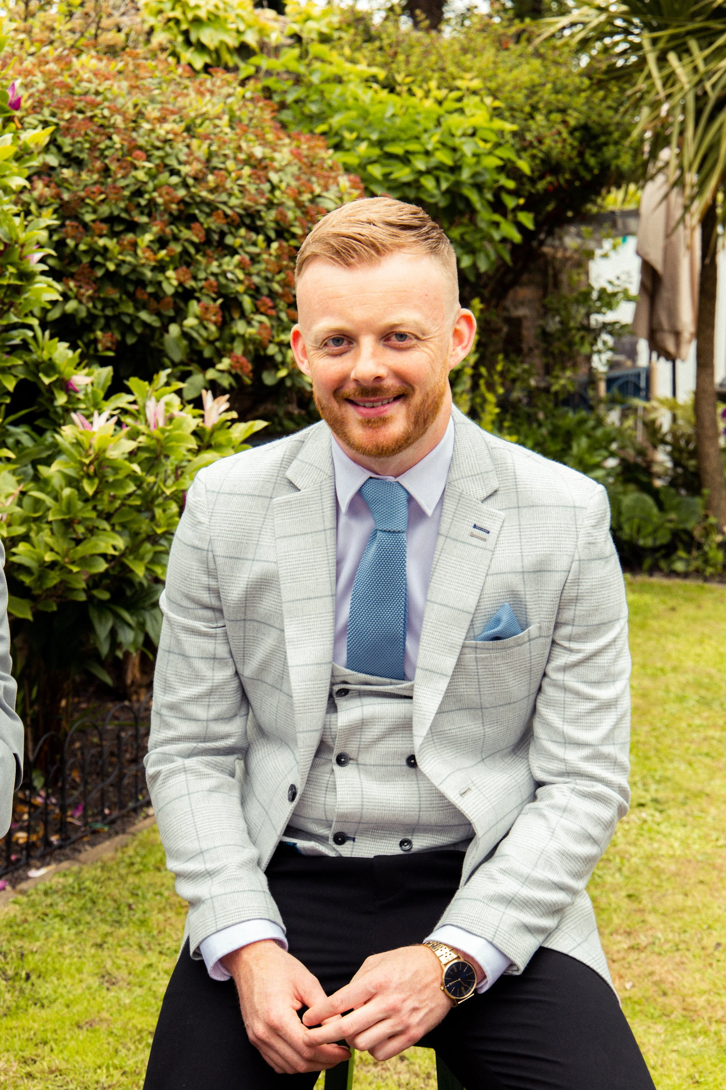 A man wearing a light grey suit with a blue tie and matching pocket square sitting outdoors in a garden with green bushes and trees, smiling at the camera.