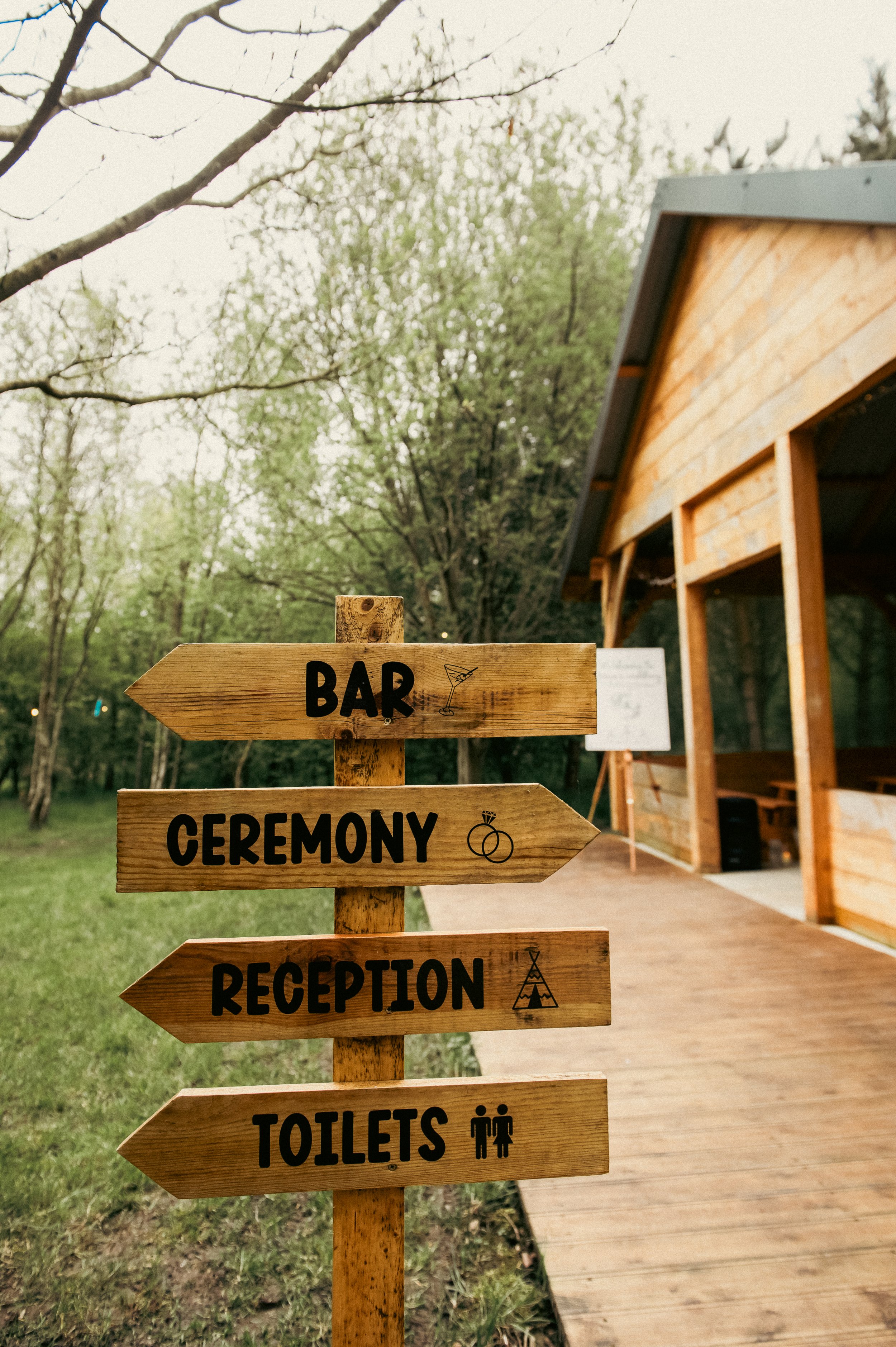 Wooden outdoor signpost with four arrows pointing to the left, labeled from top to bottom: Bar, Ceremony, Reception, Toilets, with icons of a martini glass, wedding rings, a tipi tent, and male and female symbols, against a background of trees and a 