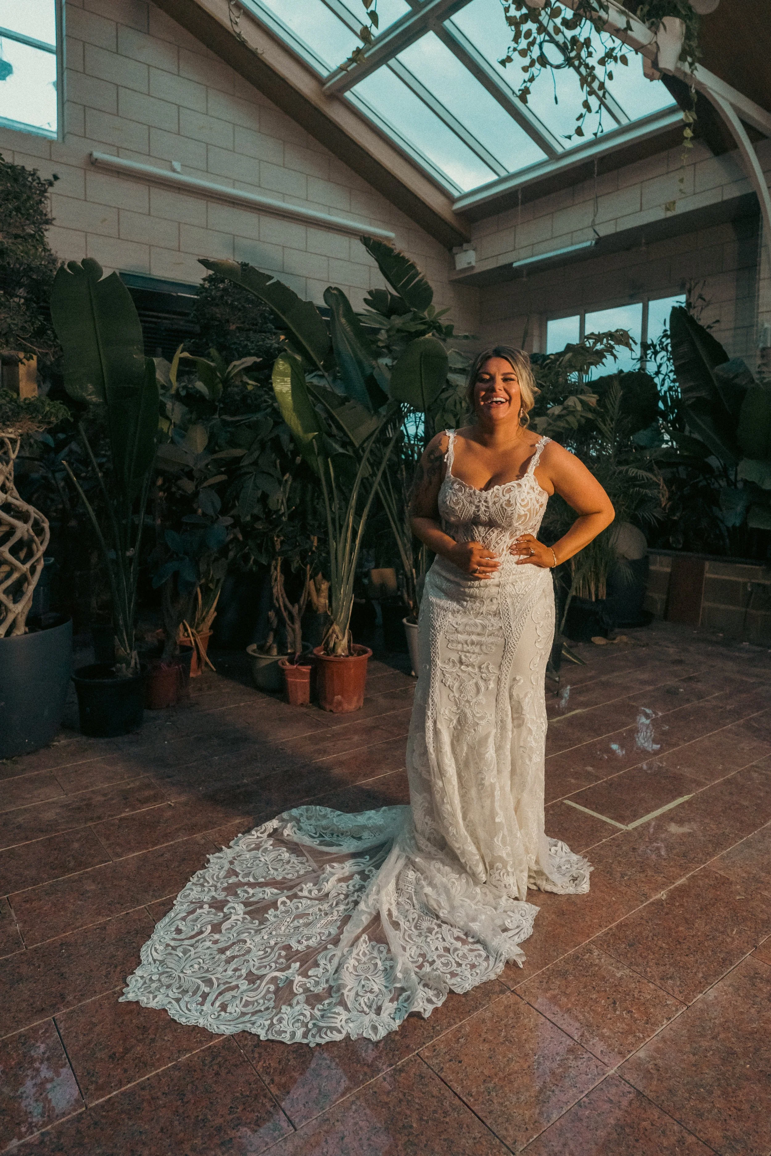 A woman in a white lace wedding dress standing indoors surrounded by lush green plants, smiling with one hand on her hip.