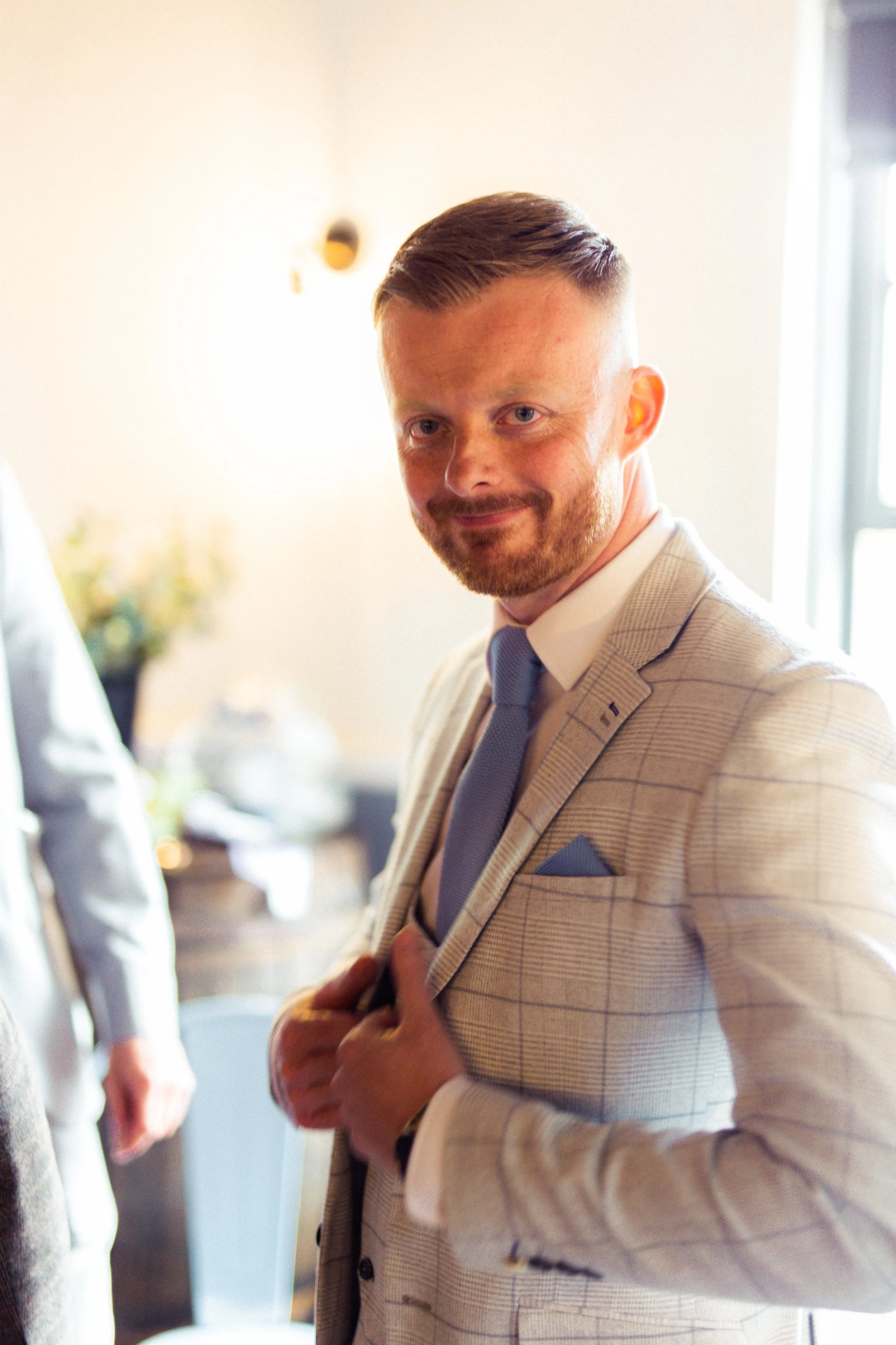 A man with short hair and a beard wearing a light-colored checkered suit, blue tie, and pocket square, smiling inside a bright room.
