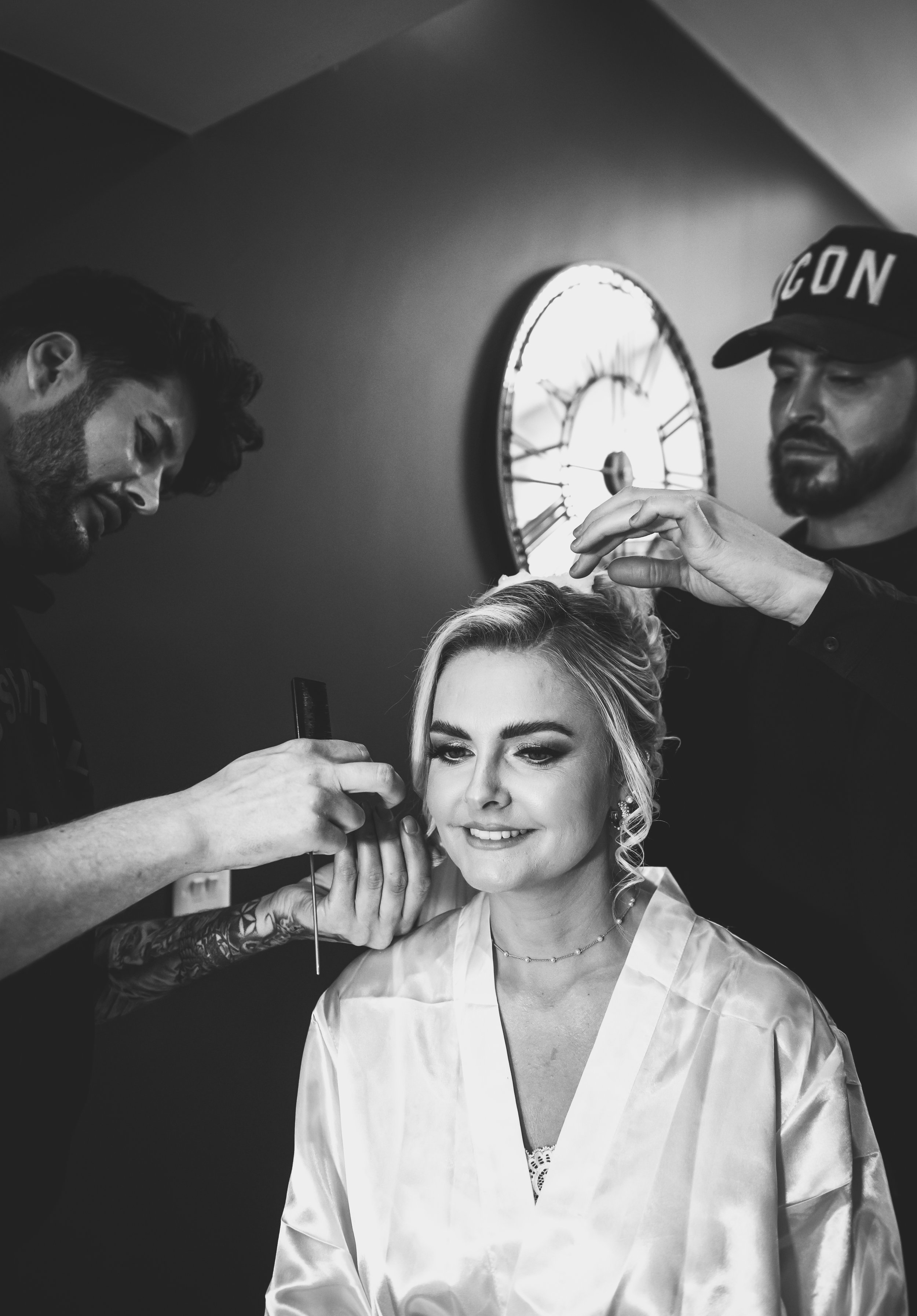 A woman with light hair sits in a salon chair, smiling as two stylists apply makeup and adjust her hair, with a large clock on the wall behind them.