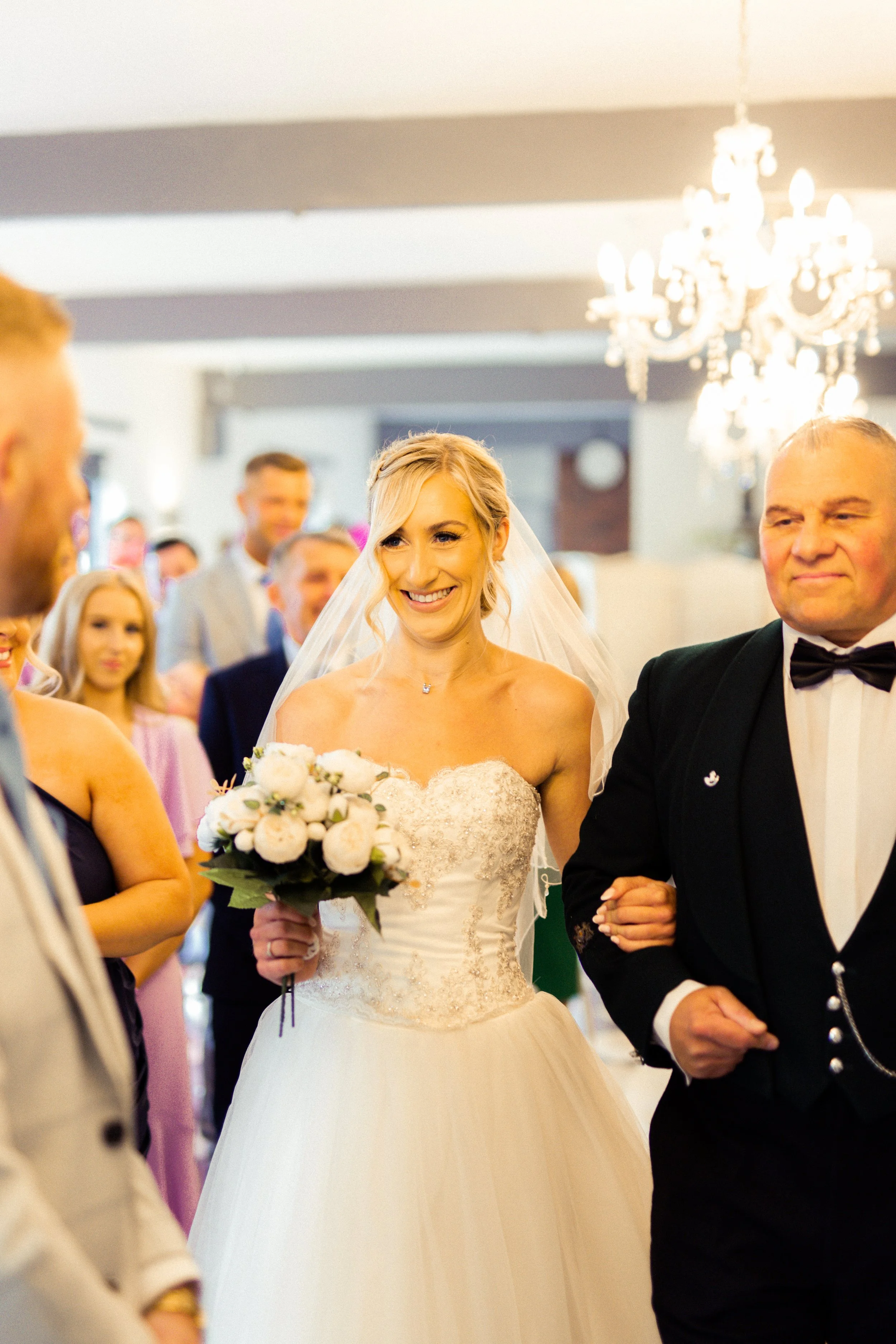 Bride holding a bouquet of white flowers, smiling, walking with a man in a tuxedo during a wedding ceremony indoors with chandelier lighting.