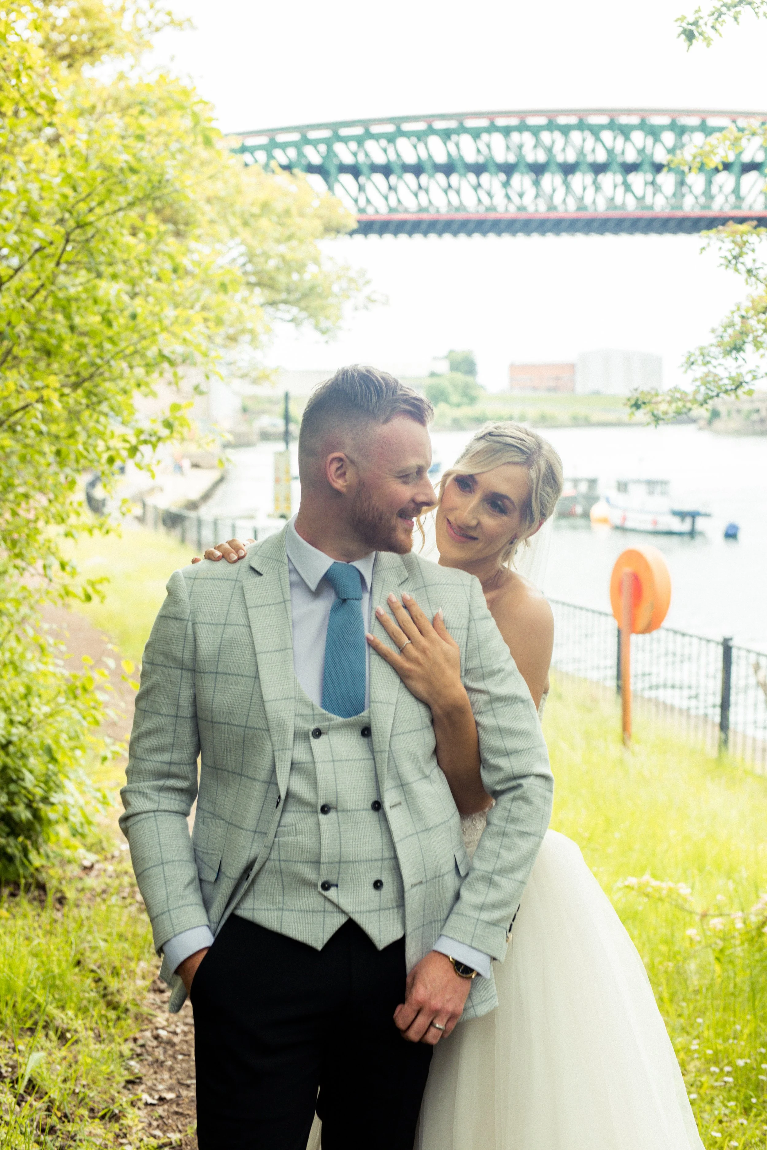 A newlywed couple, a man in a gray suit and a woman in a white wedding dress, standing by a river with boats, with a bridge in the background and green trees around them, smiling and sharing a moment.