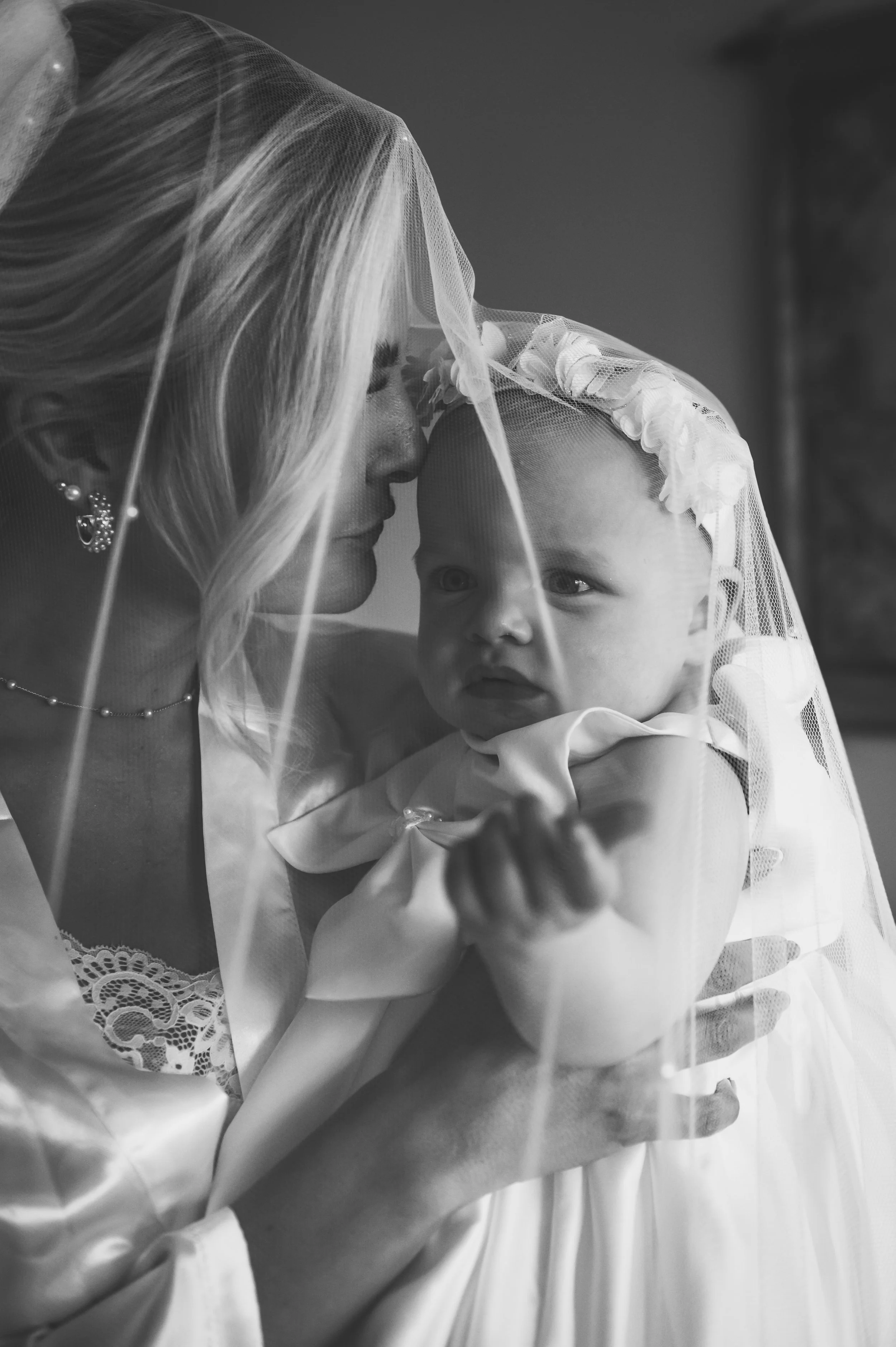 A woman, likely a bride, holding a young baby girl under a wedding veil, sharing an intimate moment indoors.