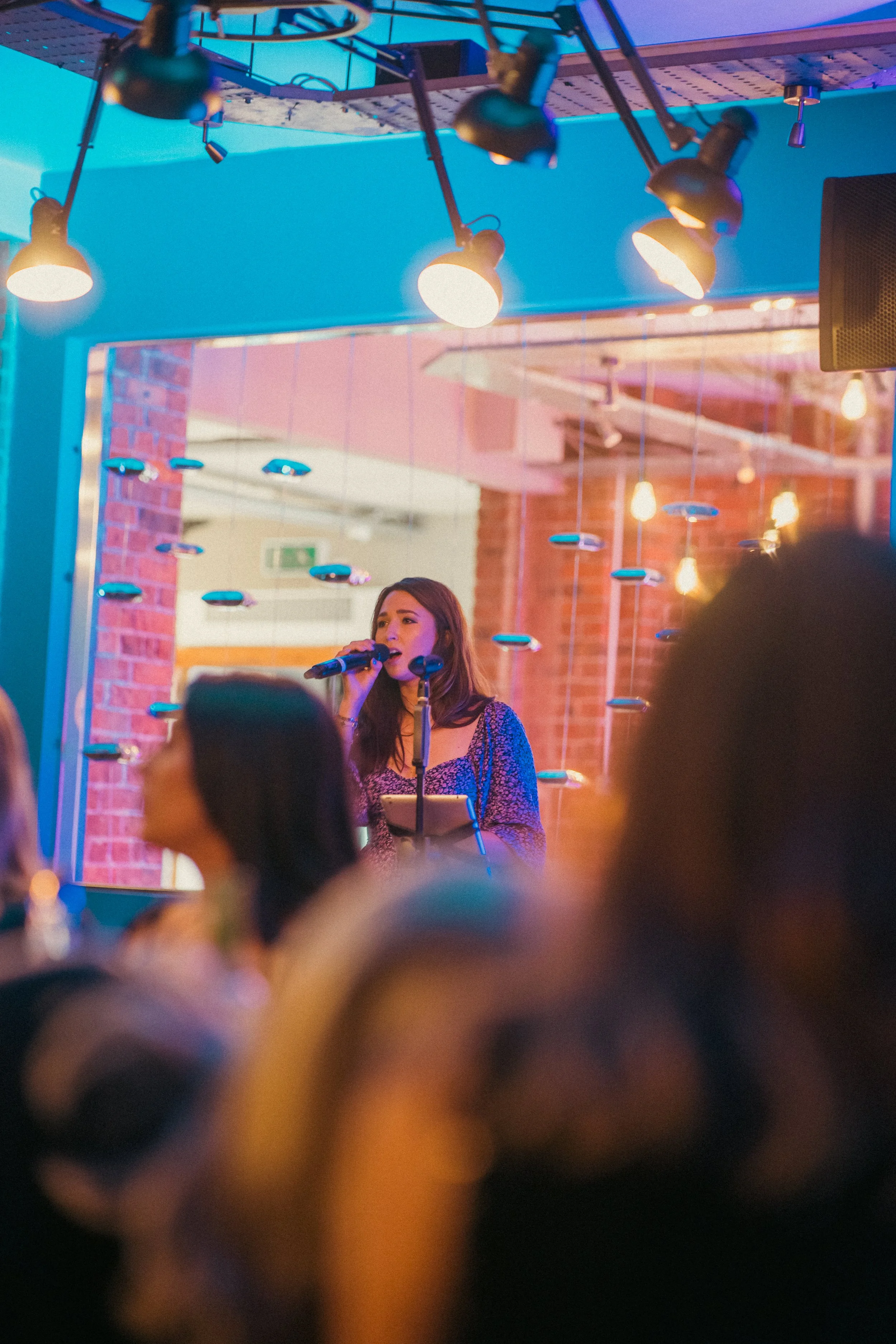 Female singer performing with microphone in a colorful, illuminated venue with hanging lightbulbs and a brick wall background.