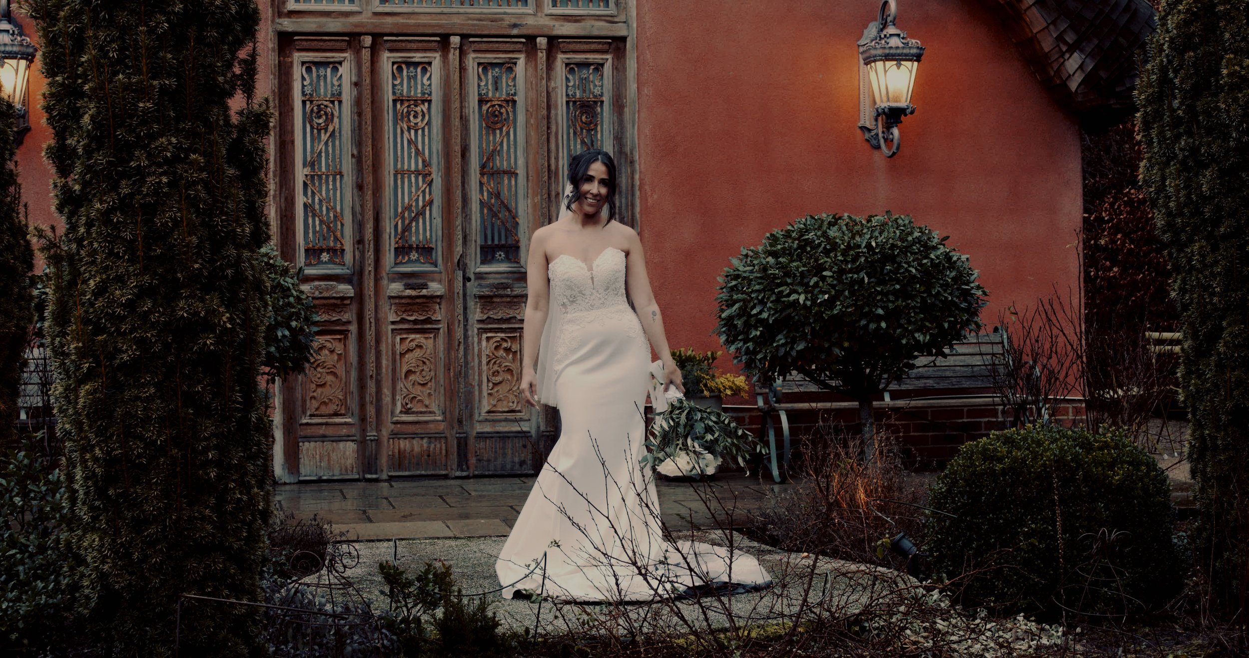 A woman in a white wedding dress standing outdoors near a rustic wooden door, with a warm lantern on the wall, surrounded by lush bushes and a small garden.
