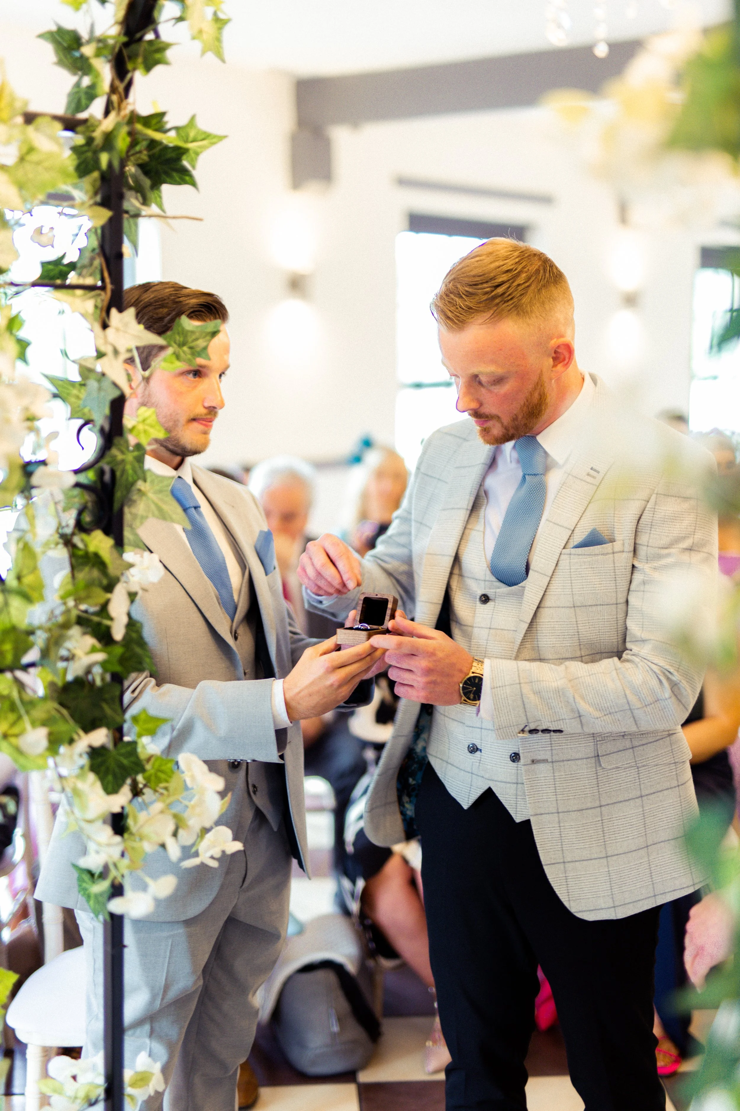 Two men in suits exchanging rings during a wedding ceremony, with guests seated in the background and floral decorations nearby.