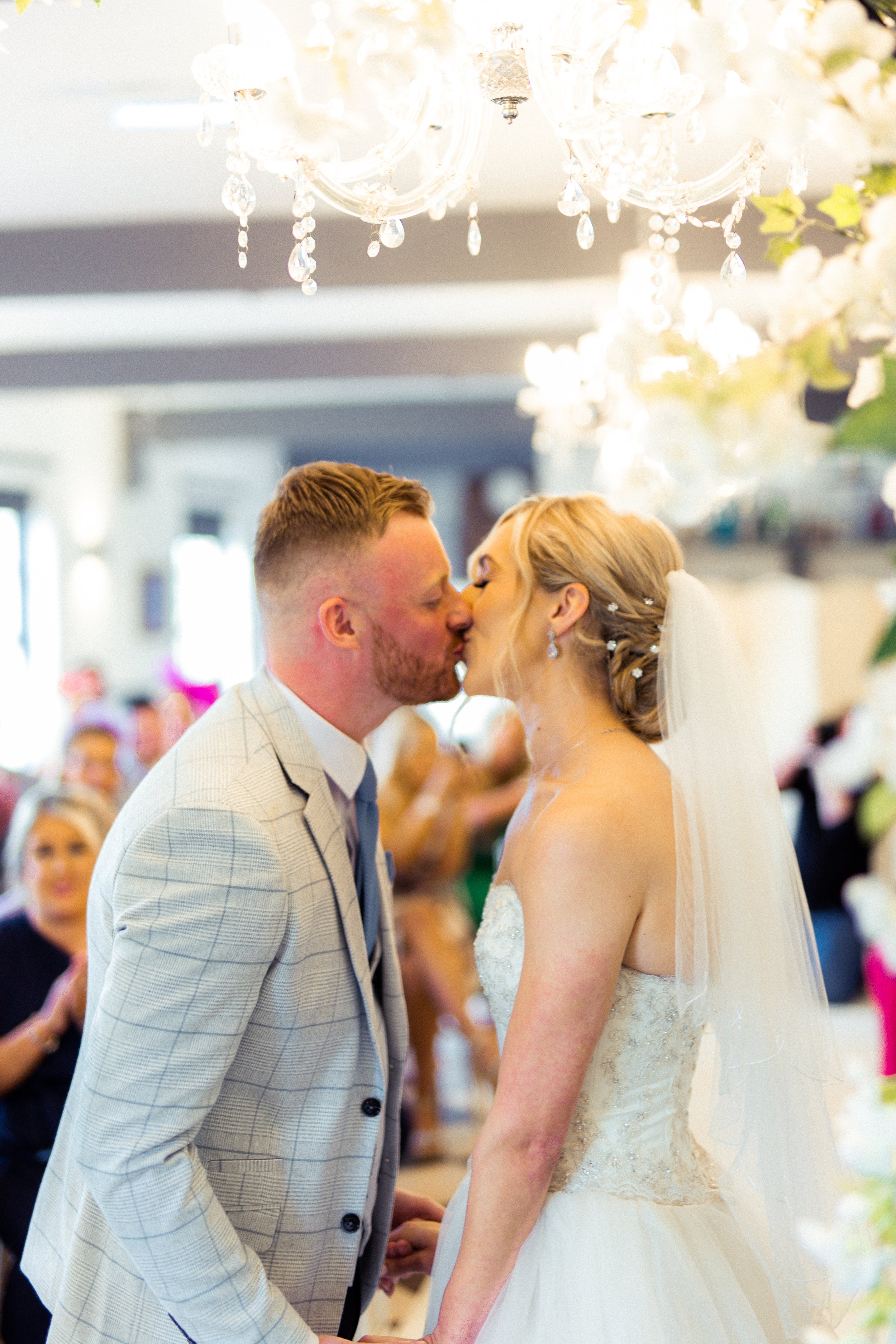 A bride and groom share a kiss at their wedding ceremony, with guests and elegant decorations including a chandelier and white flowers in the background.