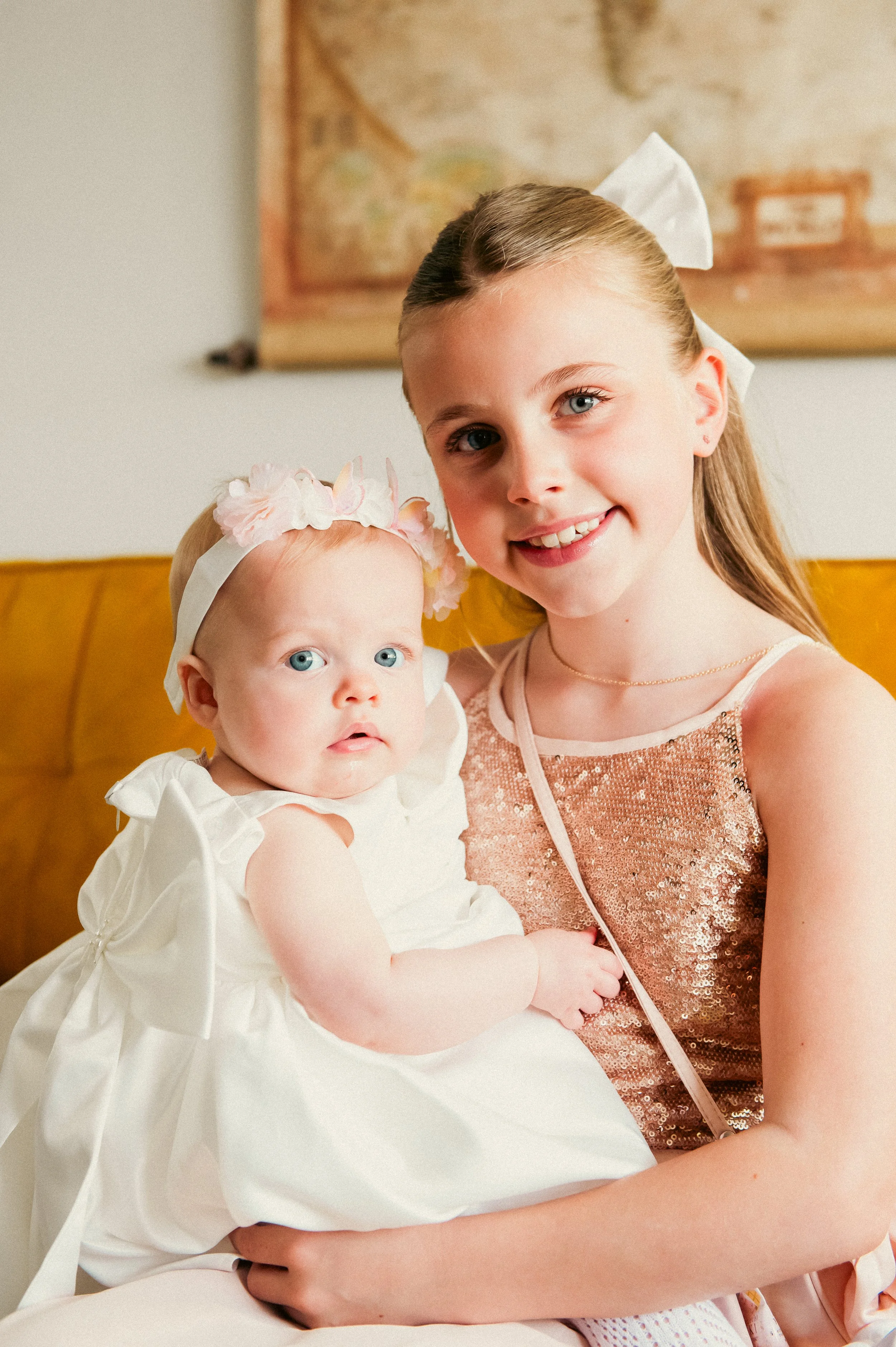 A young girl with long blonde hair, wearing a pink sequined dress and a white bow in her hair, holding a baby girl with light blonde hair, blue eyes, wearing a white dress and a floral headband, sitting on a yellow couch with a map in the background.