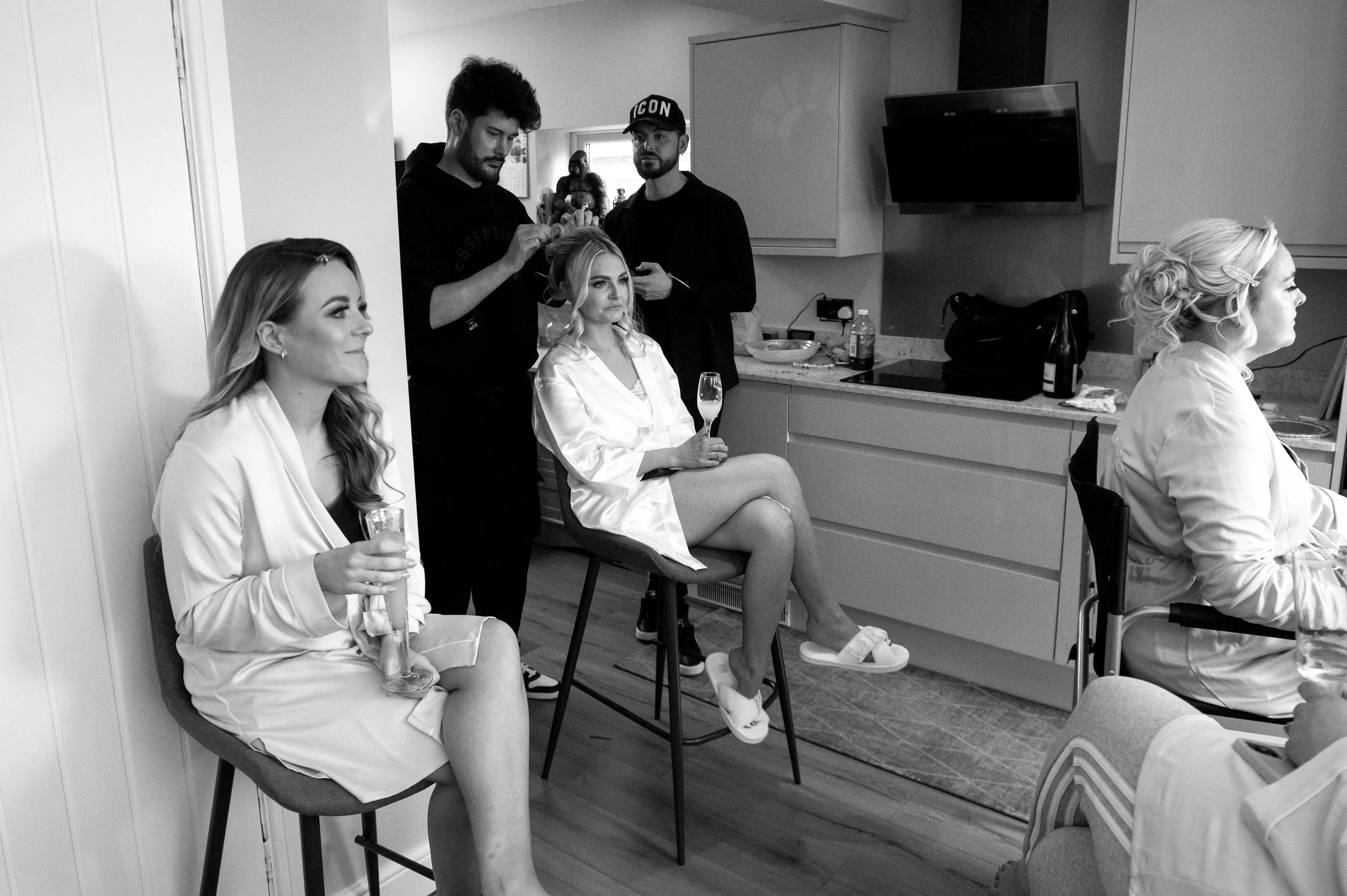 Women with satin robes sitting in a kitchen, getting ready with hairstylists and makeup artists for a special occasion.