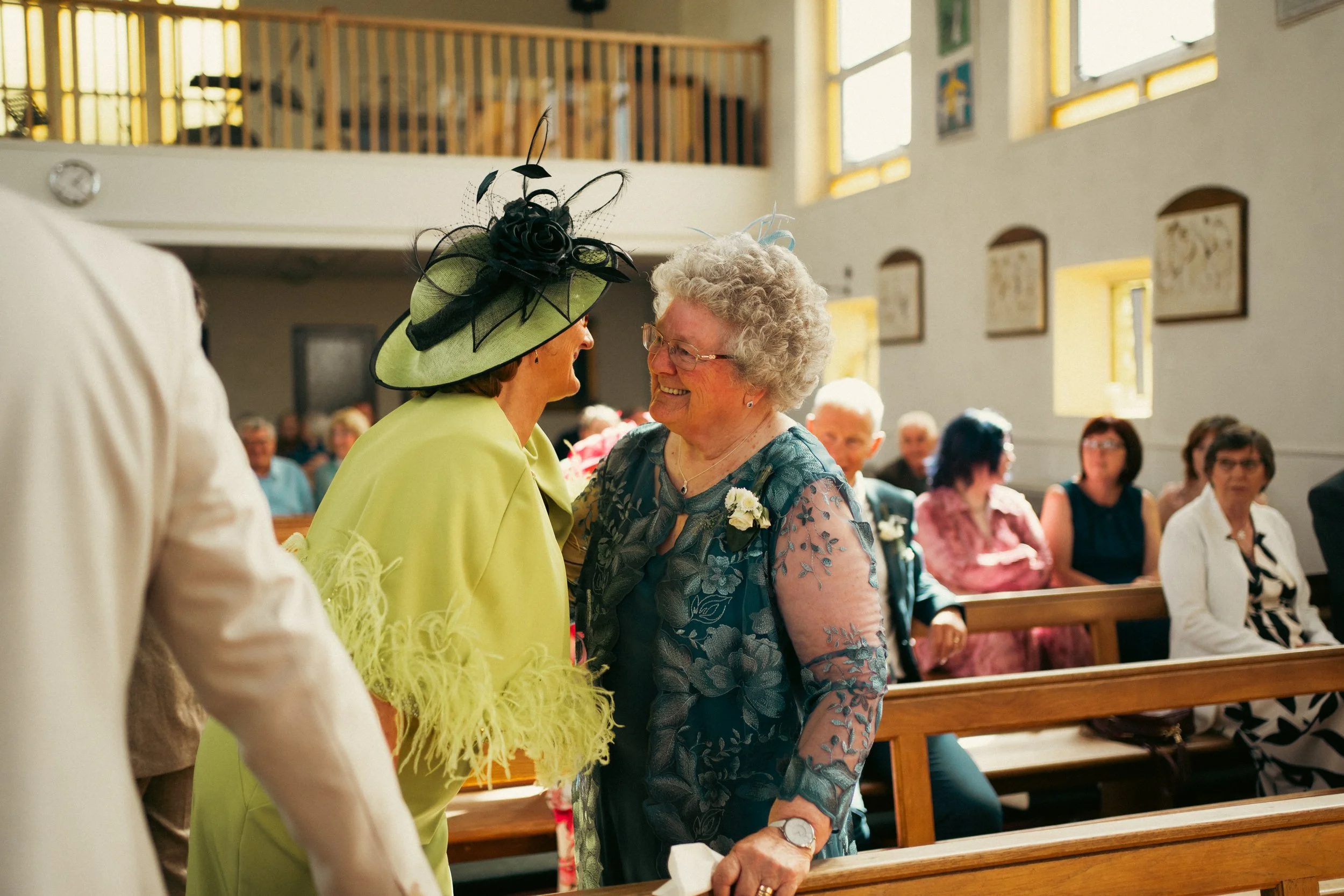 Two women greeting each other warmly in a church, one in a lime green dress and large matching hat, the other in a blue dress with floral embroidery, surrounded by seated guests.
