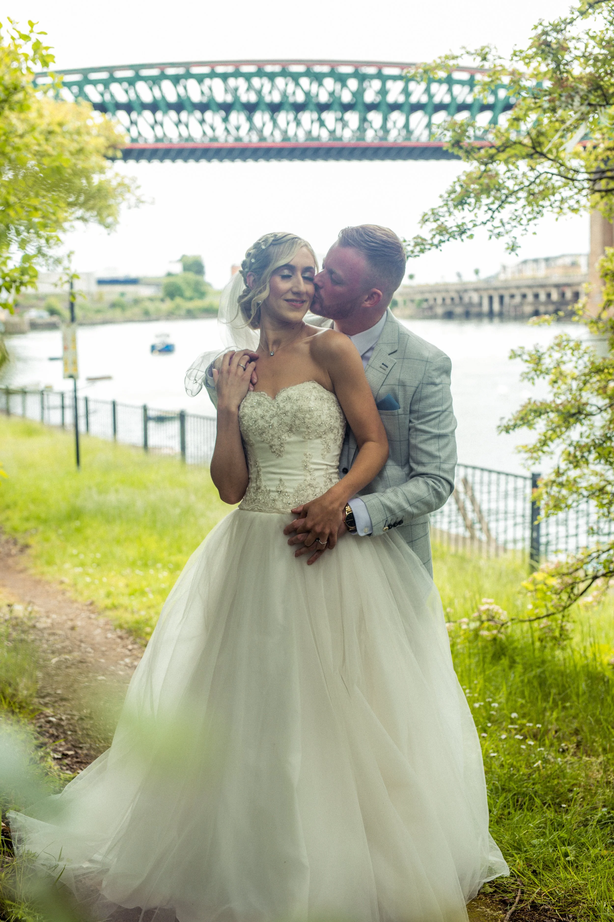 A bride and groom standing together outdoors near a river, with a bridge in the background. The bride is wearing a strapless wedding dress, and the groom is in a light gray suit. The groom is kissing the bride on the cheek as they embrace.