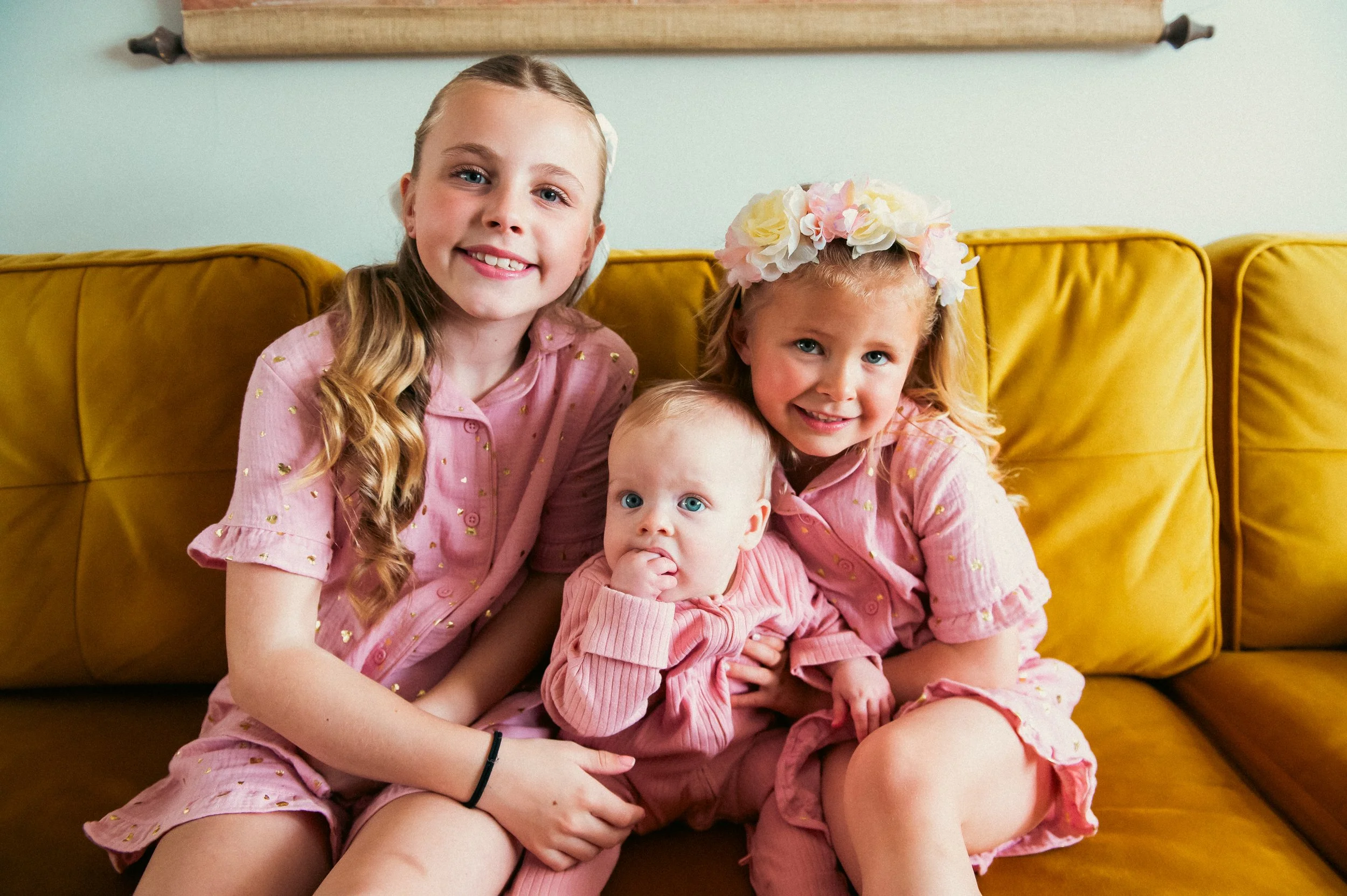 Three young girls sitting on a mustard yellow couch. One is an older girl with long, wavy blonde hair and a pink dress with gold accents. The second is a girl with curly blonde hair wearing a pink dress with a floral headband. The youngest is a baby 