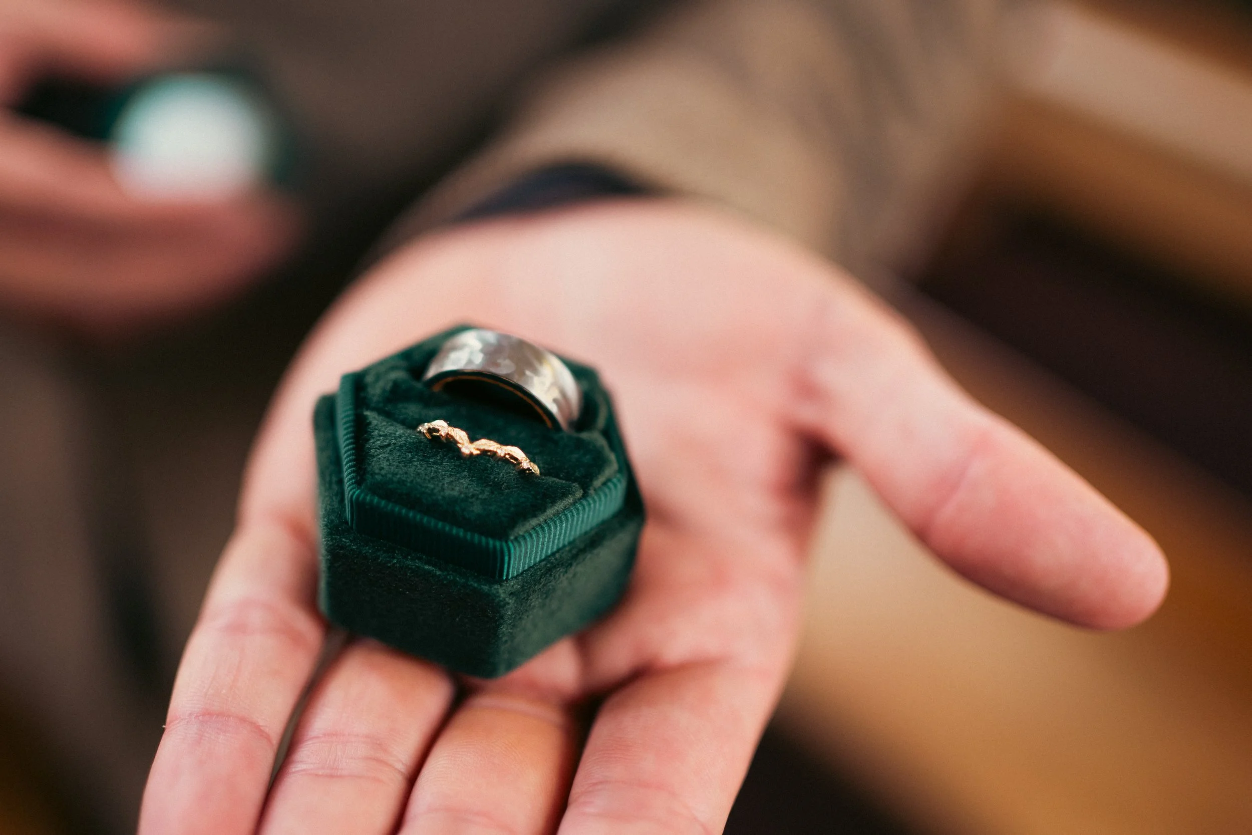 Close-up of a person's hand holding a green velvet ring box with two rings inside: a silver wedding band and a gold wedding band, with a blurred background.