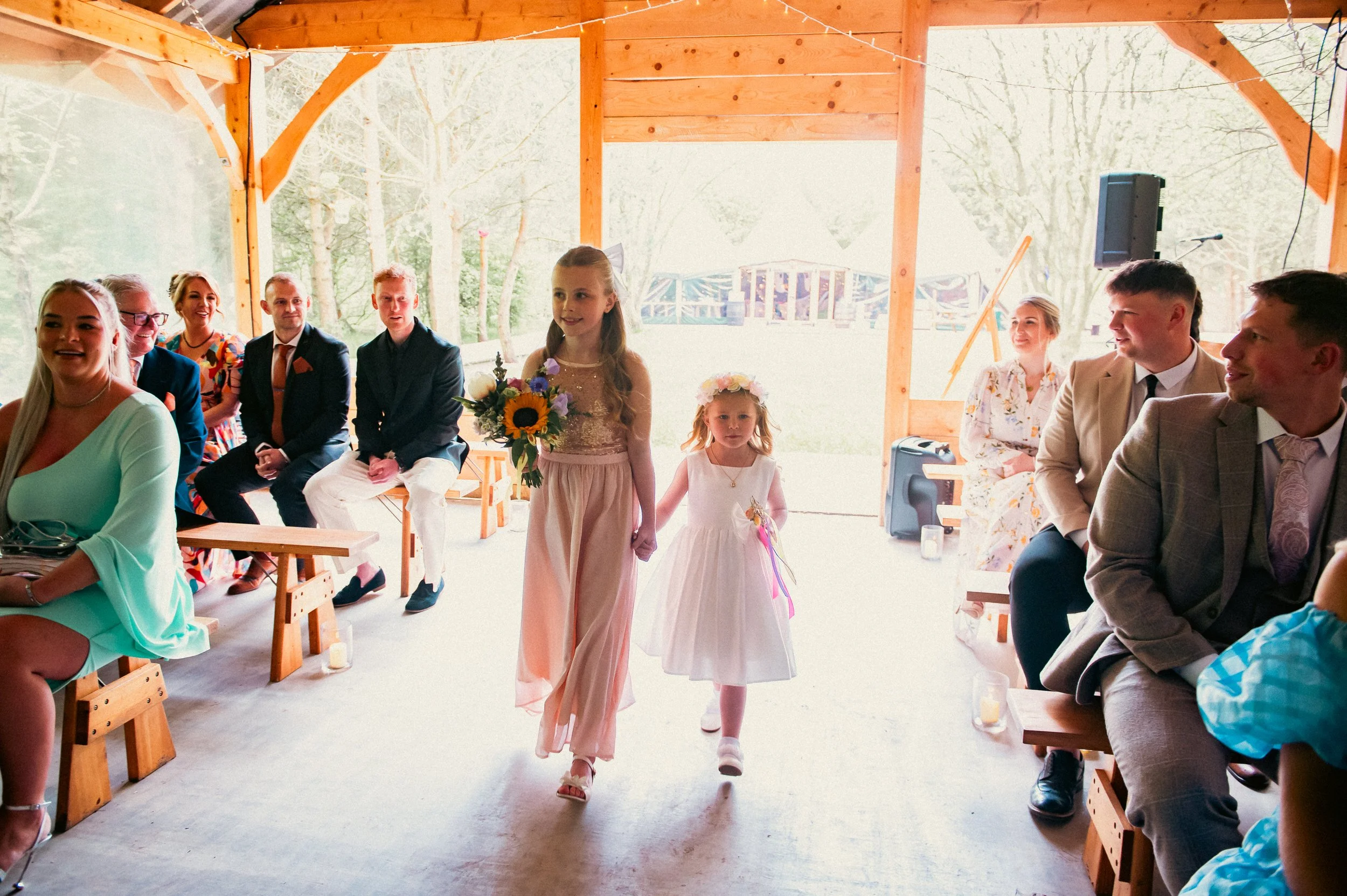 Two young girls in white dresses walking down the aisle at a wedding ceremony, holding hands, with guests seated on wooden benches on either side, indoors with a wooden structure and open back view of trees.