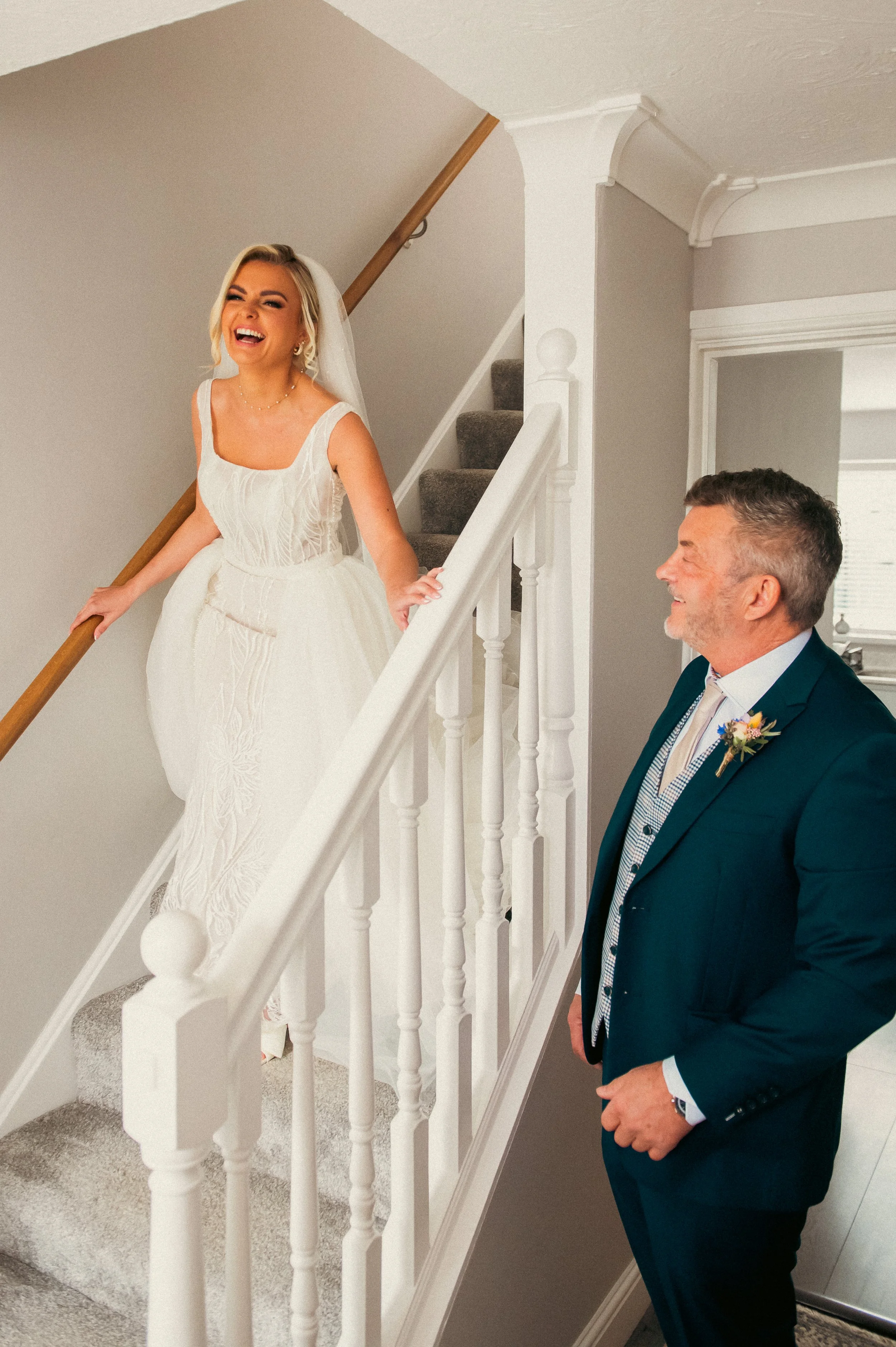 A bride in a white wedding dress smiling and laughing while standing on the stairs, with a man in a dark suit looking up at her.