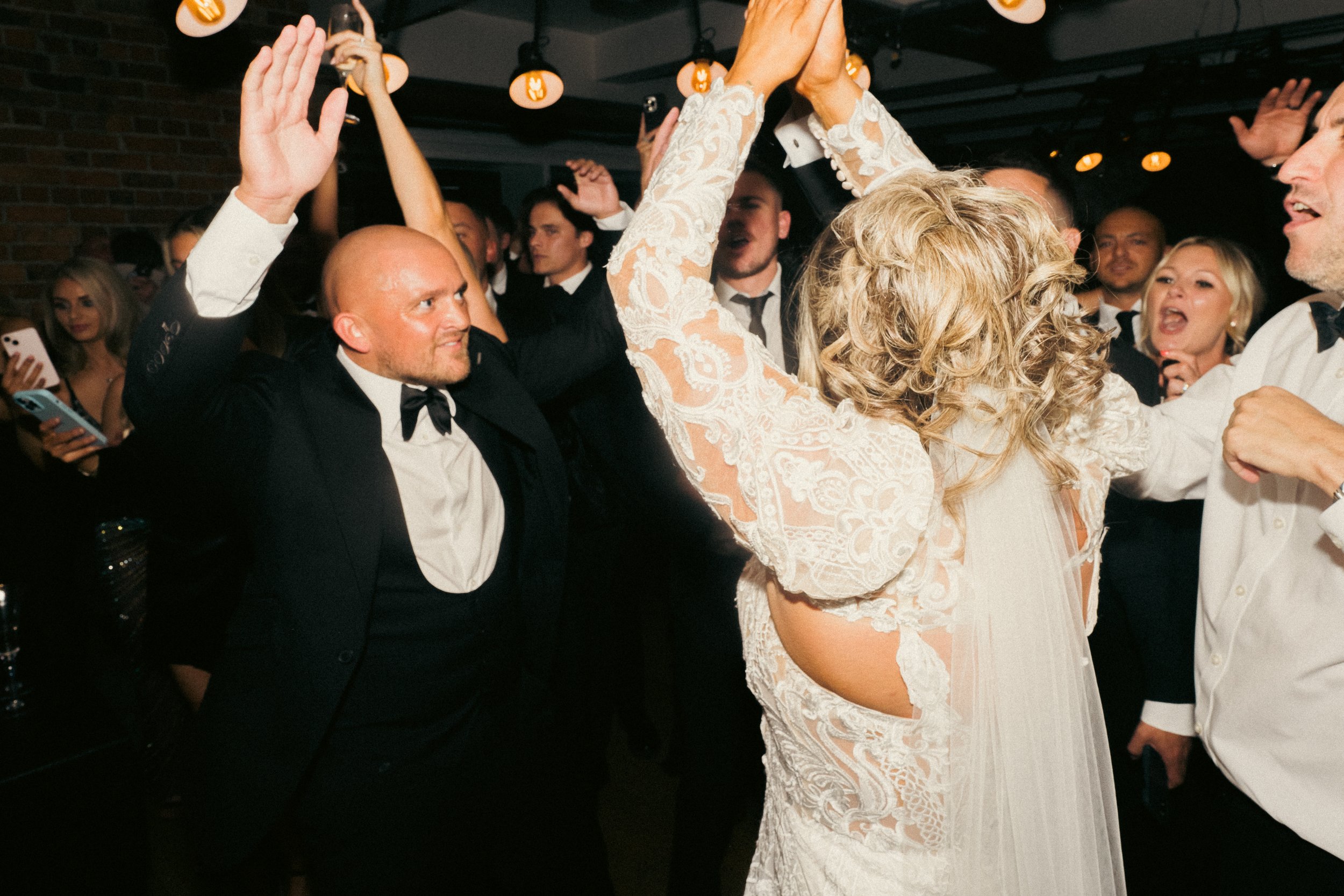 Couples dancing at a wedding reception with guests in formal attire, celebrating indoors with warm lighting.