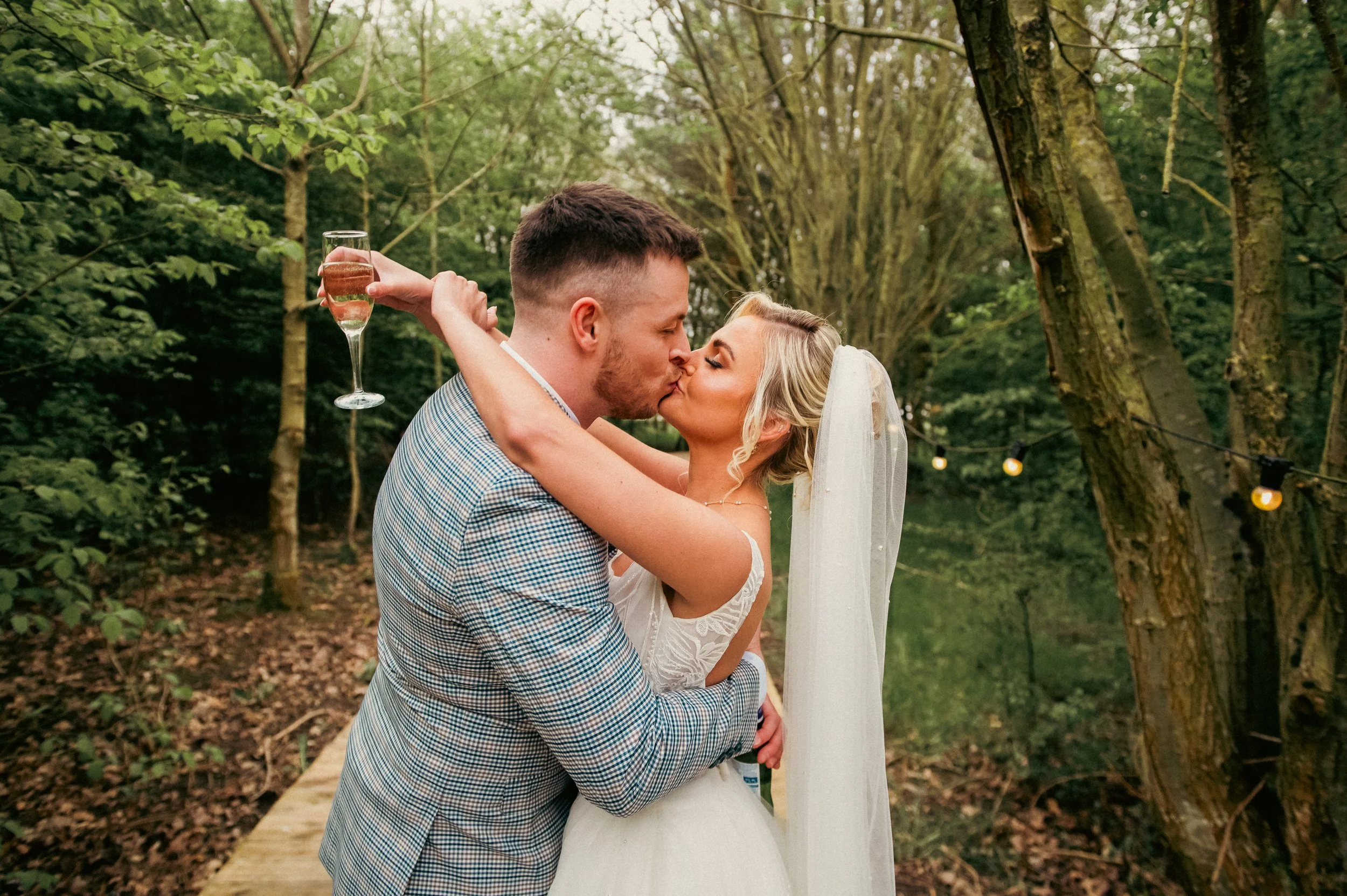 A newlywed couple sharing a kiss in a wooded outdoor setting, with string lights overhead. The groom holds a glass of champagne behind the bride as they embrace.