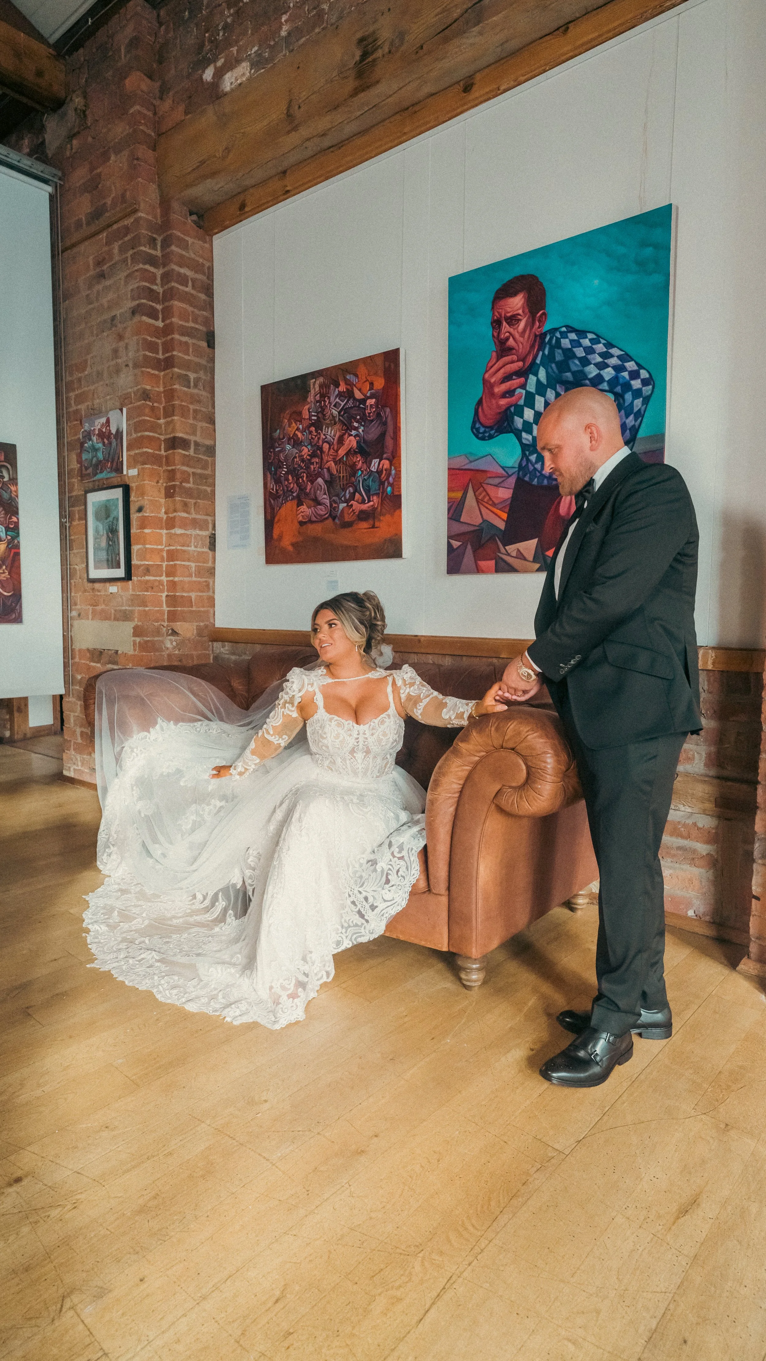 Bride sitting on a brown leather sofa in a white wedding gown, holding the hand of a man in a black tuxedo, inside an art gallery with colorful paintings on brick and white walls.