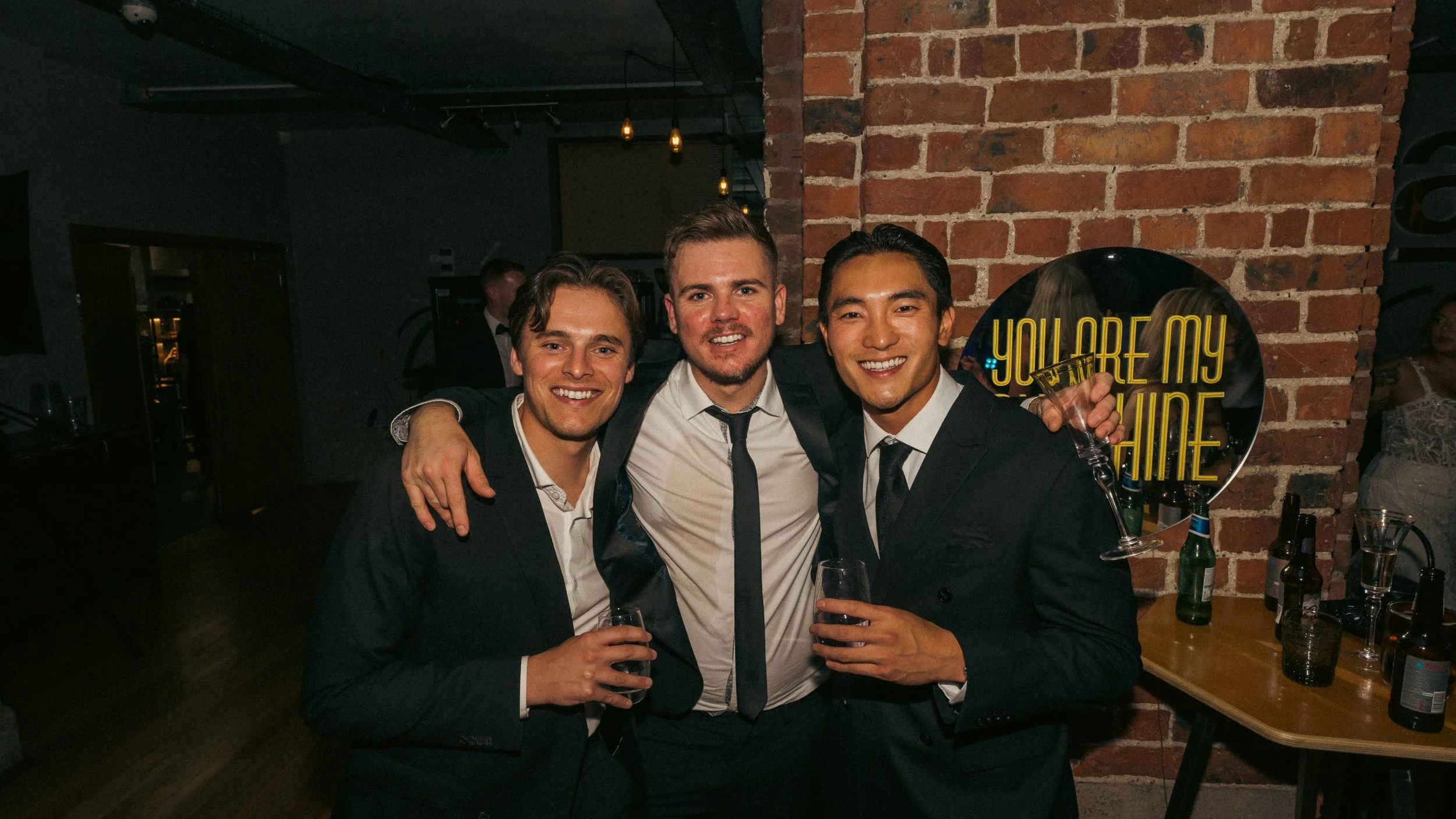 Three men in suits at a party, smiling and holding drinks, standing in front of a brick wall with a neon sign that reads 'You Are My Sunshine'.